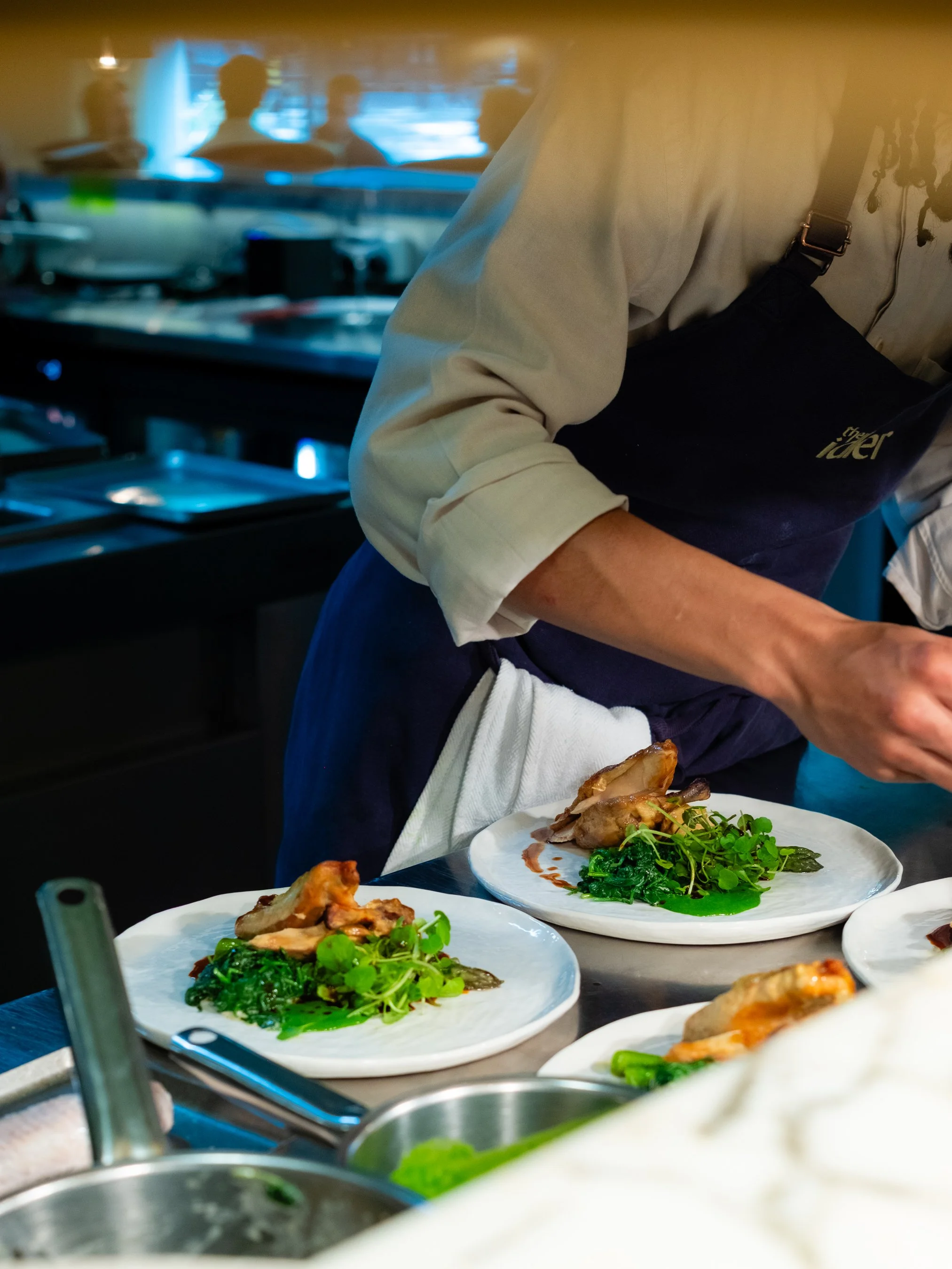 Food plates with greens and meat in a professional kitchen with a chef in the background.