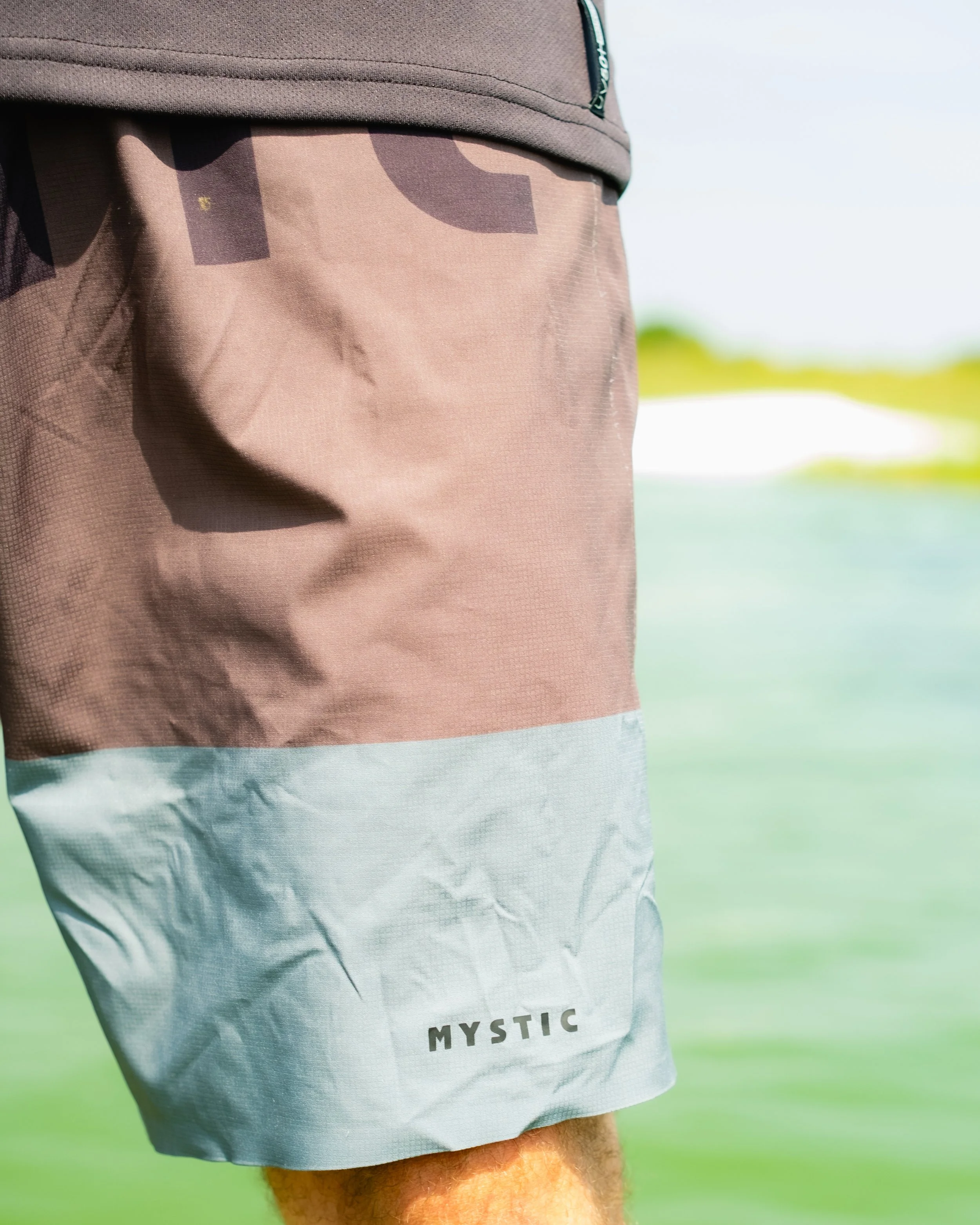 Close-up of a person wearing beige and grey Mystic brand shorts, standing outdoors near water and grass.