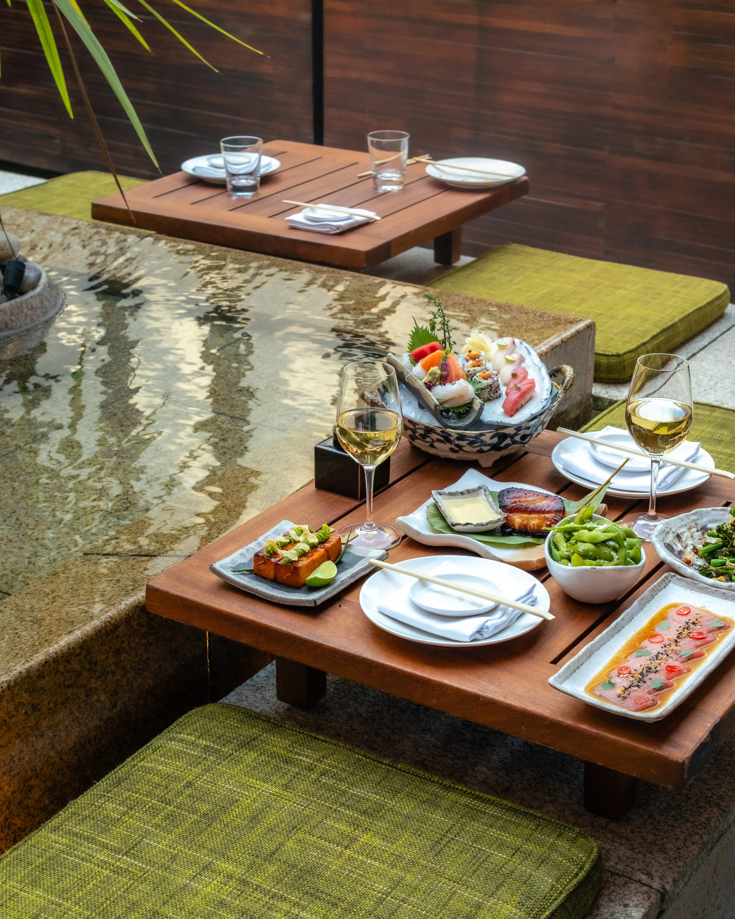 A Japanese-style outdoor dining area with a low wooden table, green cushion seats, and a variety of sashimi and other dishes, along with glasses of white wine, under a tree with green leaves.