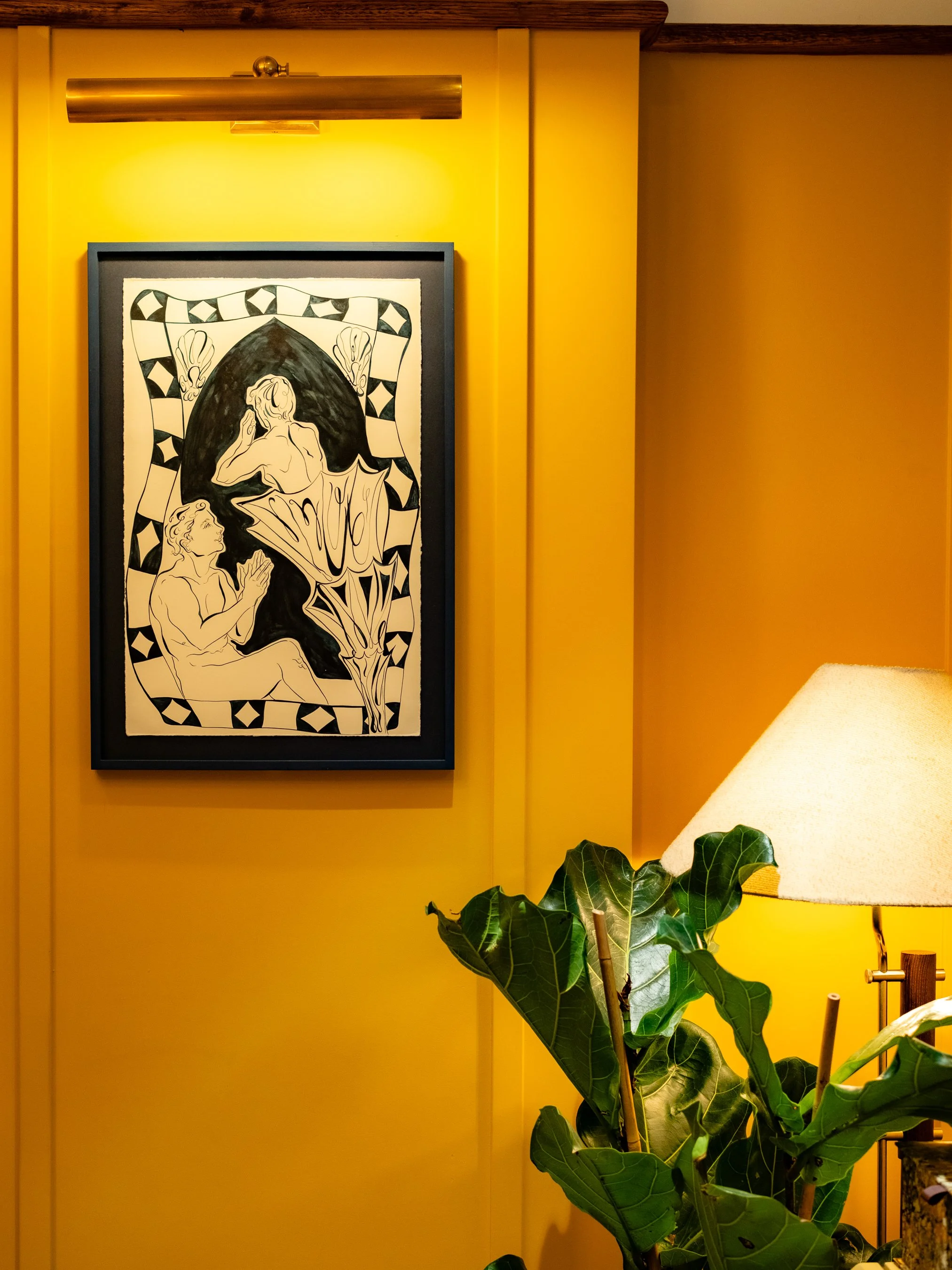 Painting of two people, one sitting and one with a hand to their face, in black and white, framed by a black frame on a yellow wall, with a lamp above and a large green leafy plant and a beige lampshade in the foreground.