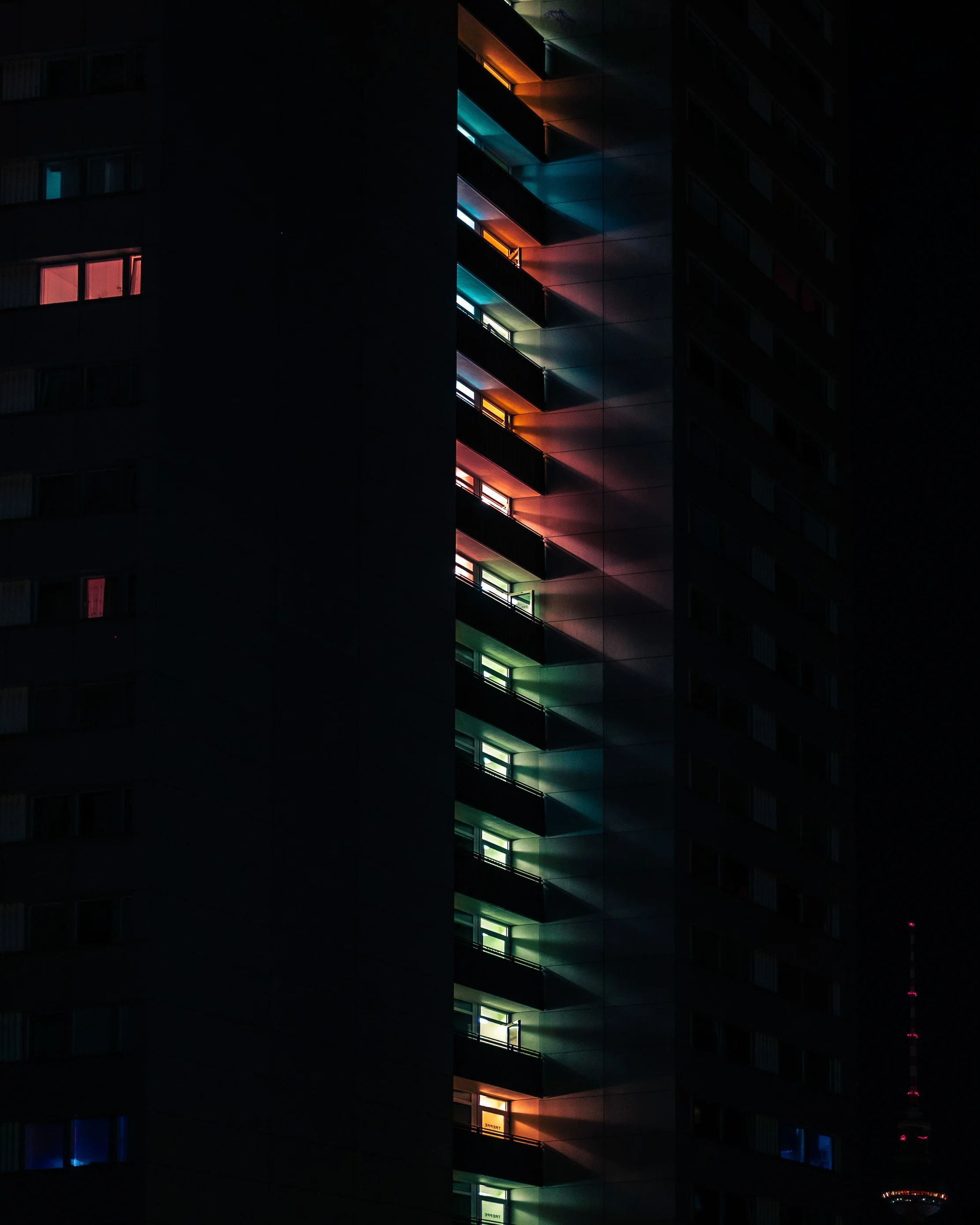 Nighttime view of a high-rise building with illuminated windows in various colors, including red, yellow, green, blue, and white, creating a vibrant pattern on the dark facade.