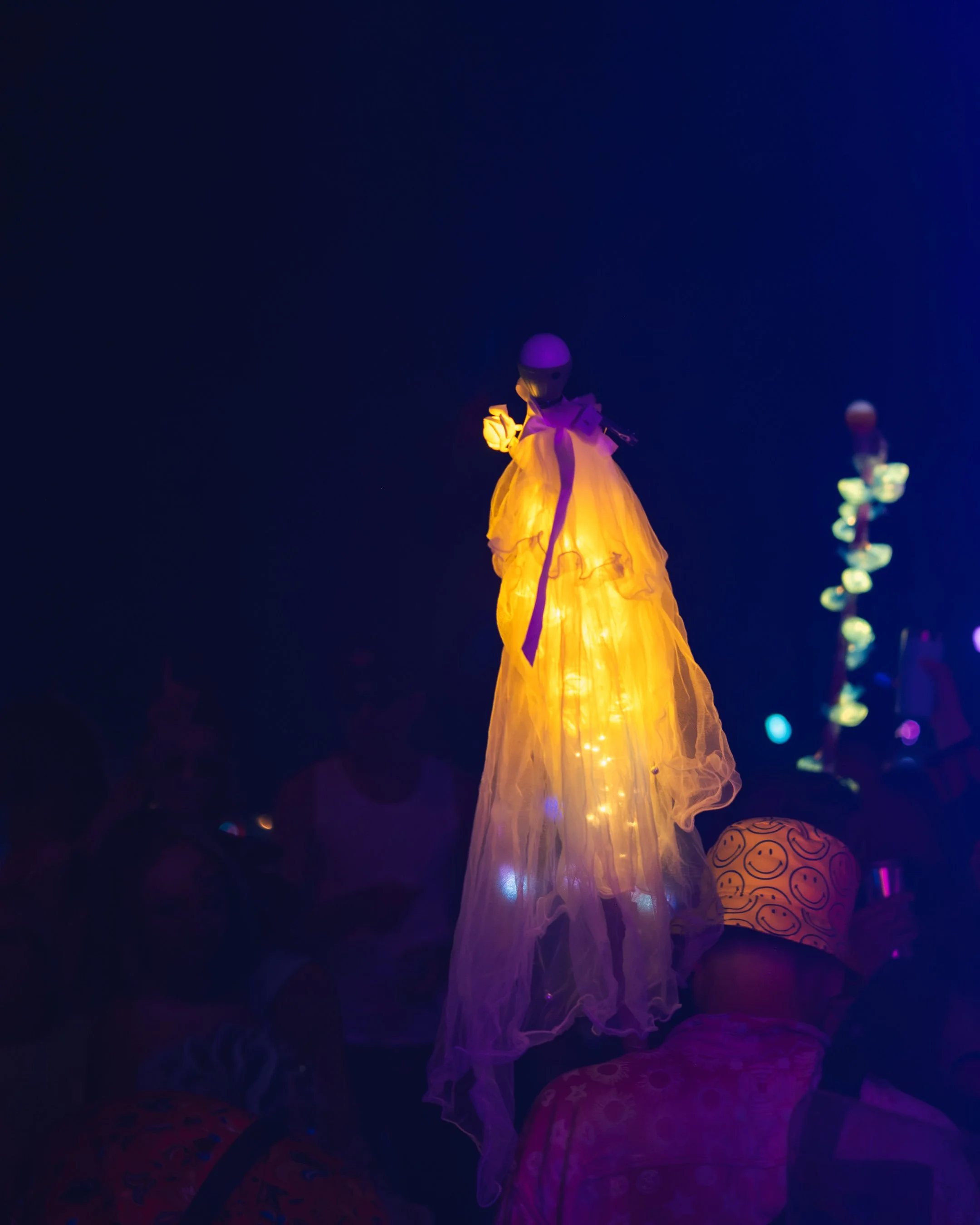 A decorative figure resembling a doll or sculpture, illuminated with warm yellow and purple lights, standing against a dark background. To the right, there is a smaller, colorful object with smiley face patterns.