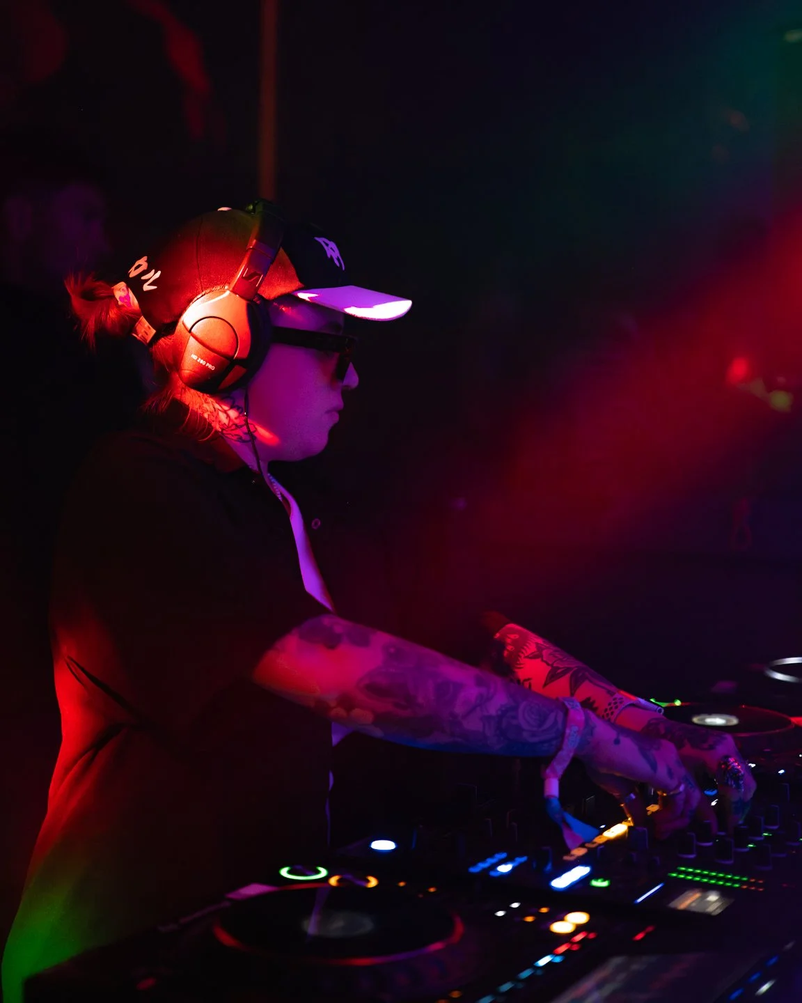 A DJ with tattoos wearing headphones, sunglasses, and a cap, playing music at a dark, colorful nightclub or concert.