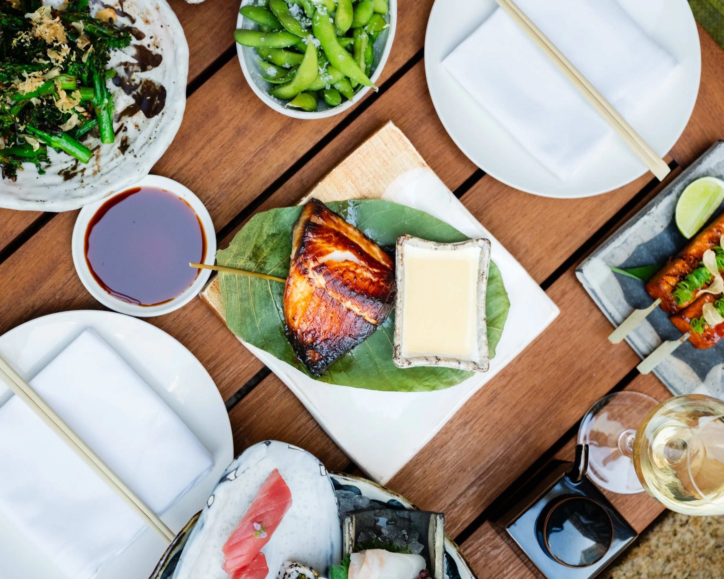 A table with various Asian dishes, including a bowl of edamame, a plate with grilled fish on a banana leaf, soy sauce, and a hot dog with sliced pink fish cake, along with empty white plates and a glass of wine.