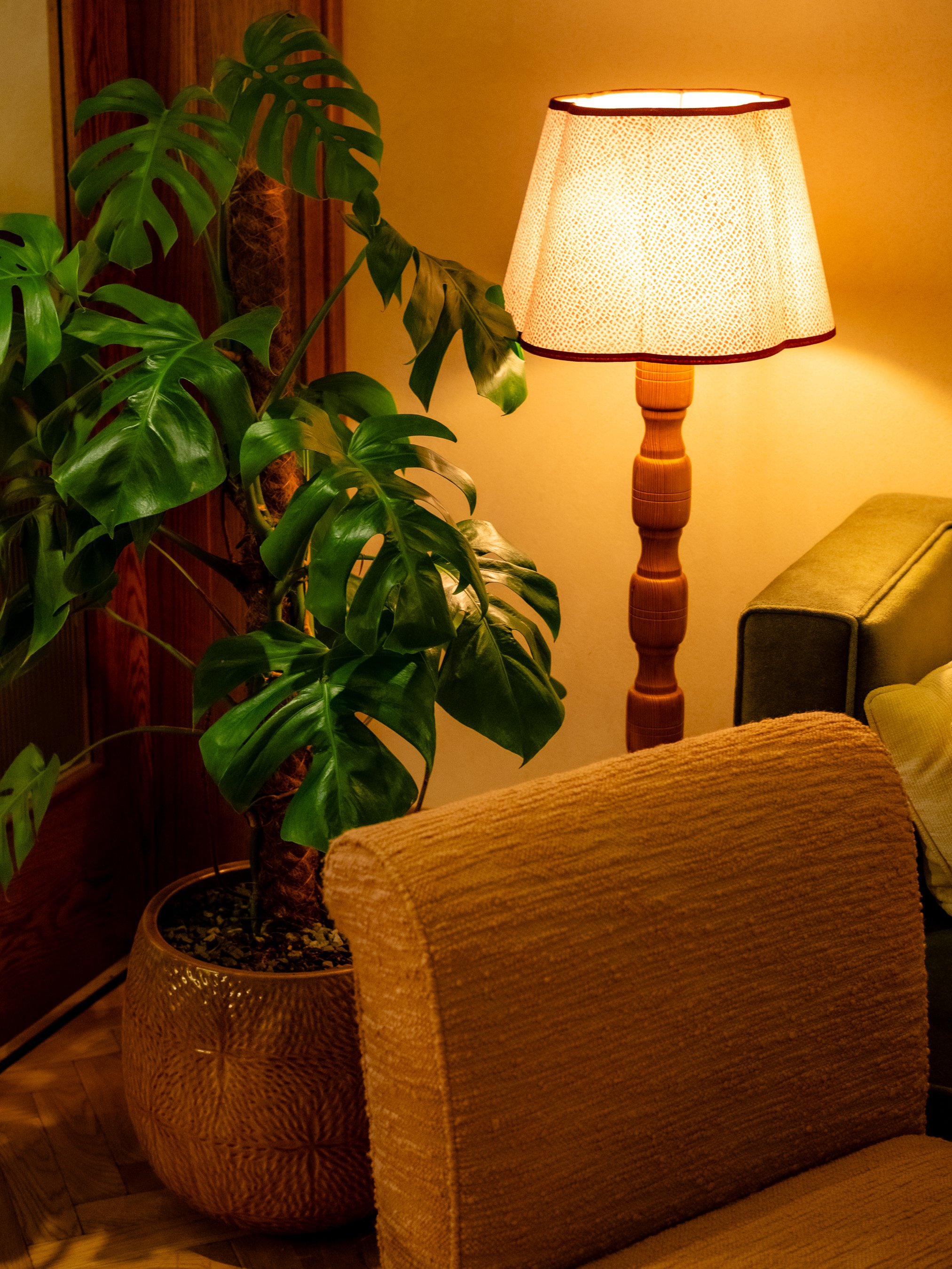 A living room corner with a tall wooden lamp, a green leafy plant, a textured beige armchair, and a woven basket.