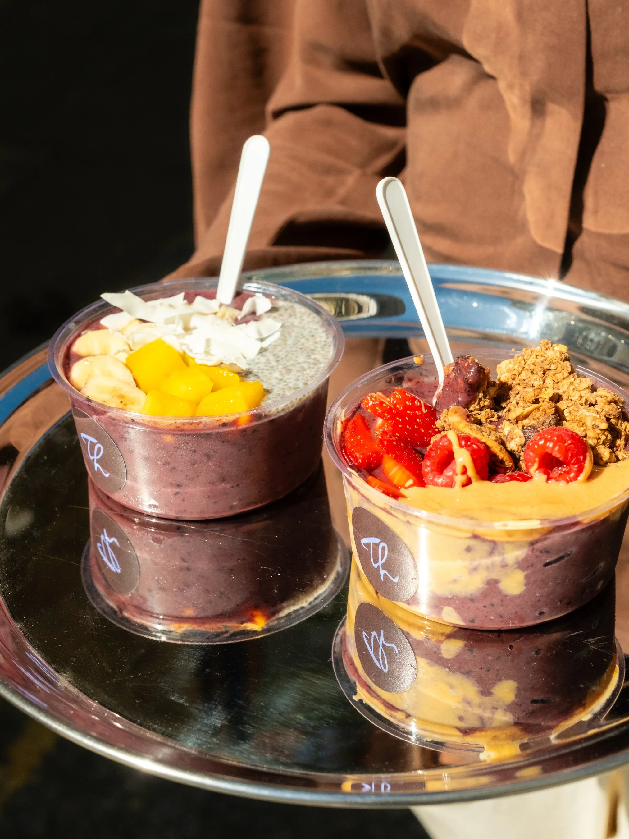 Two cups of mixed berry smoothies topped with sliced fruit, white chocolate shavings, granola, and strawberries on a reflective silver tray.