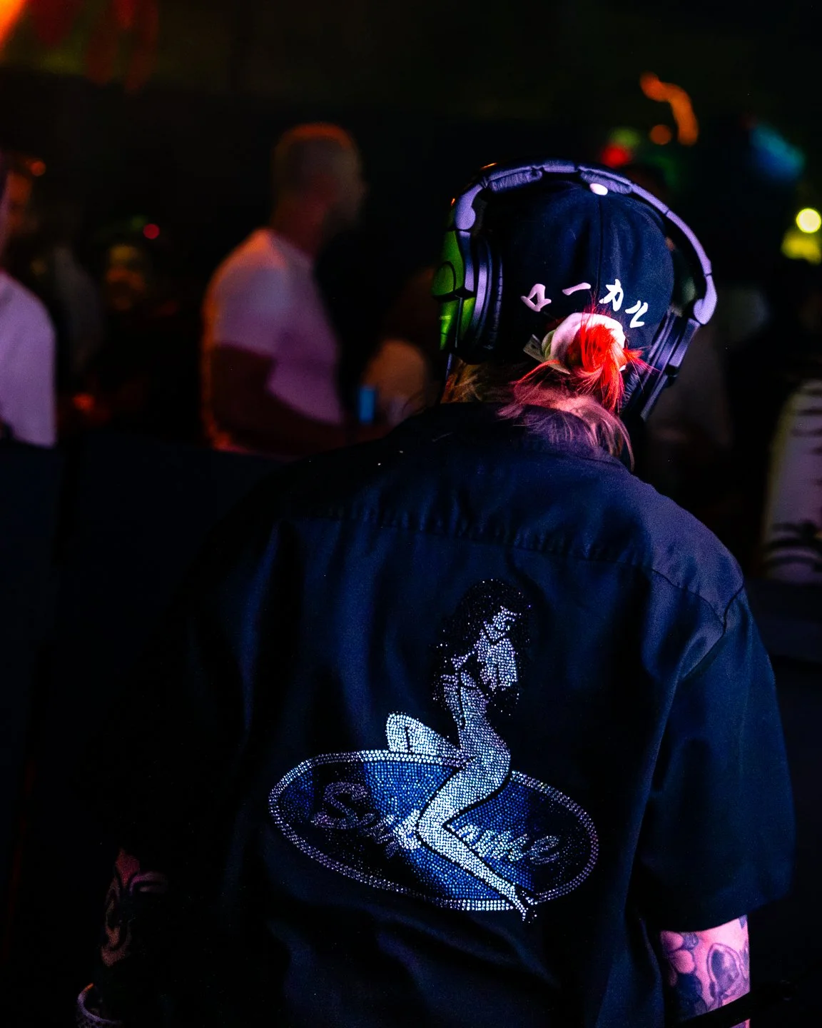 Person with pink hair wearing a black beanie and headphones, black jacket with a rhinestone logo of a mermaid and the word 'Sealine' on the back, in a dark setting with colorful lights and blurred people in the background.