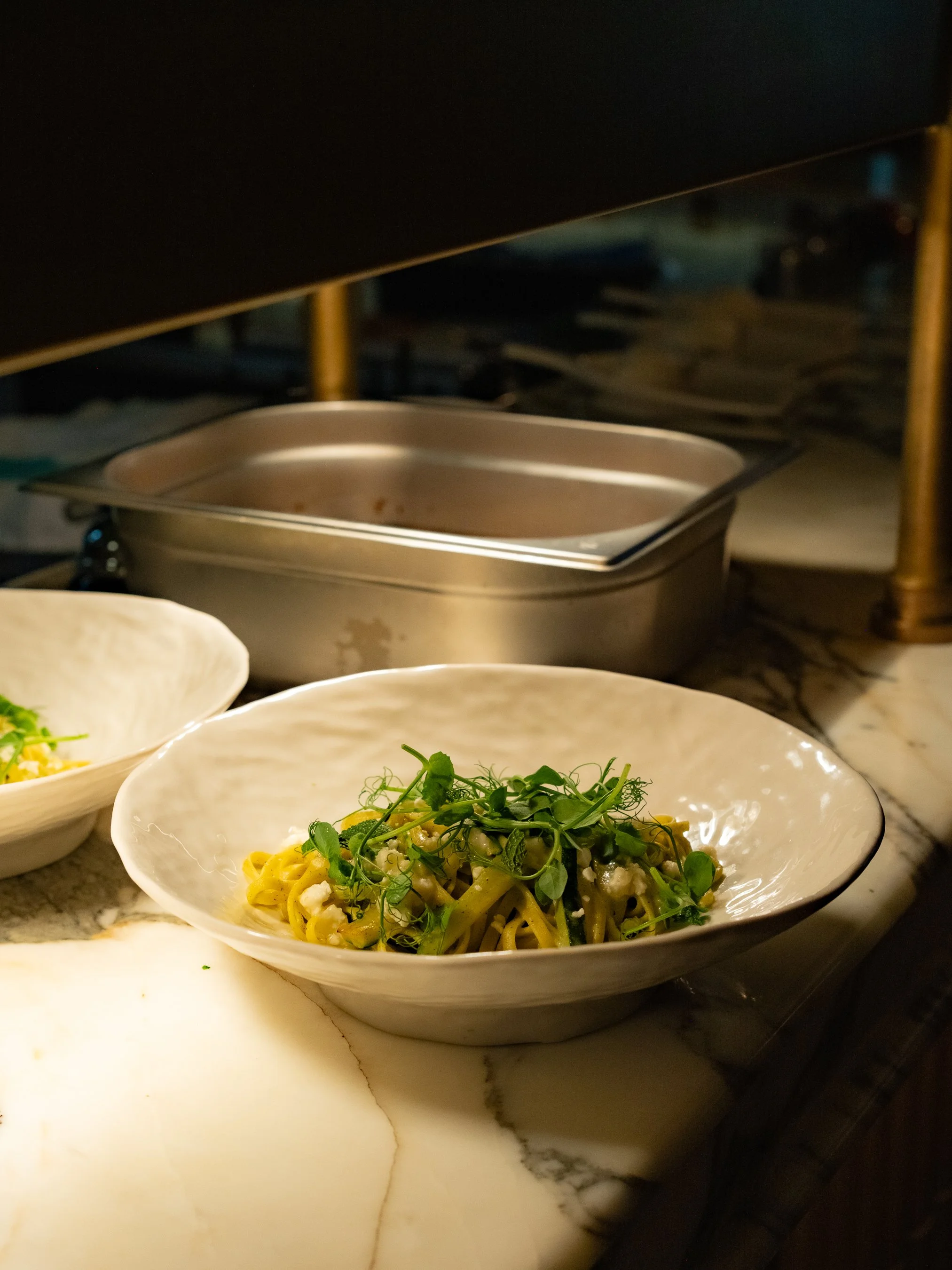 A white bowl of pasta topped with microgreens on a marble counter, with other dishes and kitchen equipment in the background.