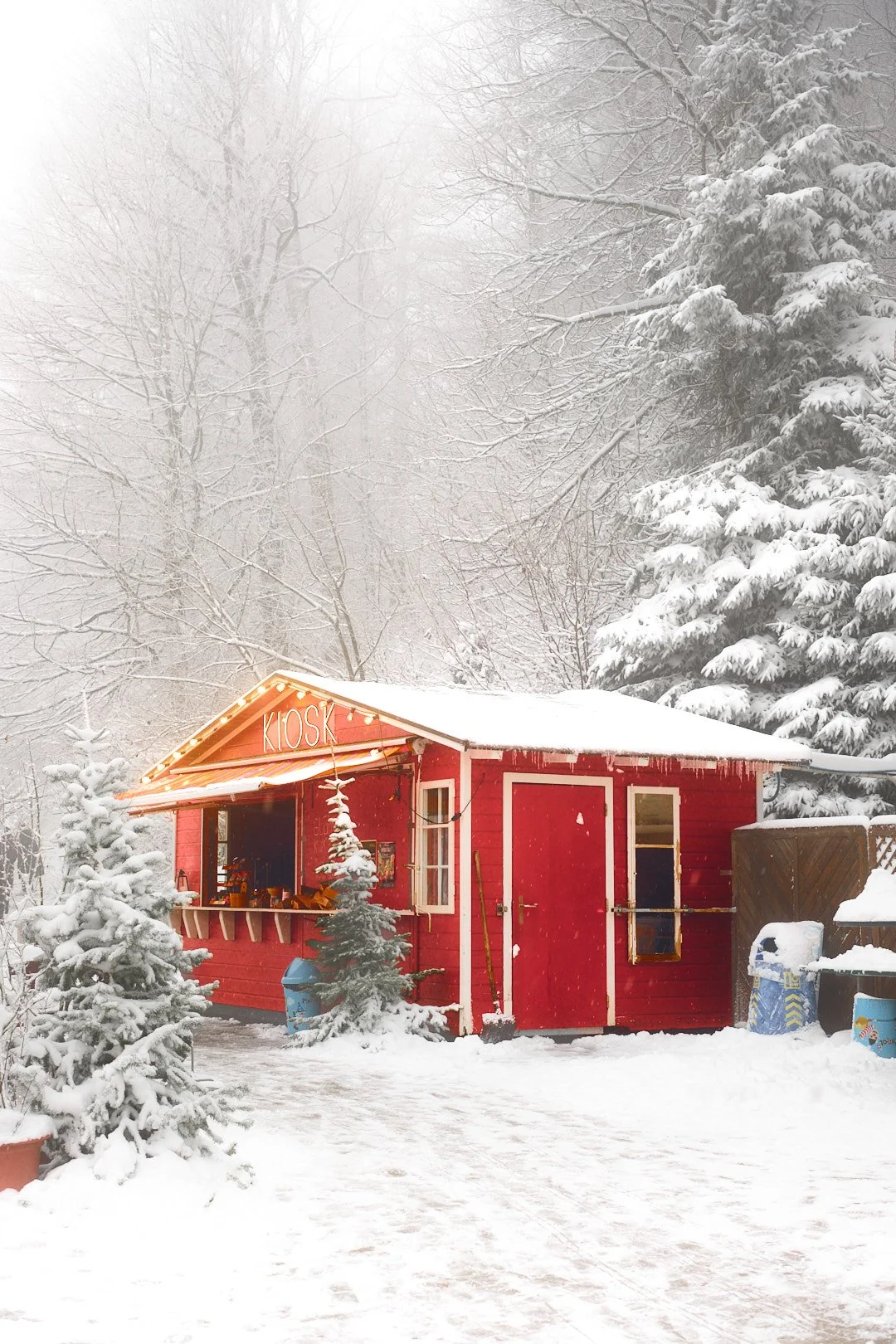 A small red kiosk with lights and a sign reading 'KIOSK' in a snowy winter landscape. Snow-covered trees surround the kiosk, and snow is on the ground and roof.