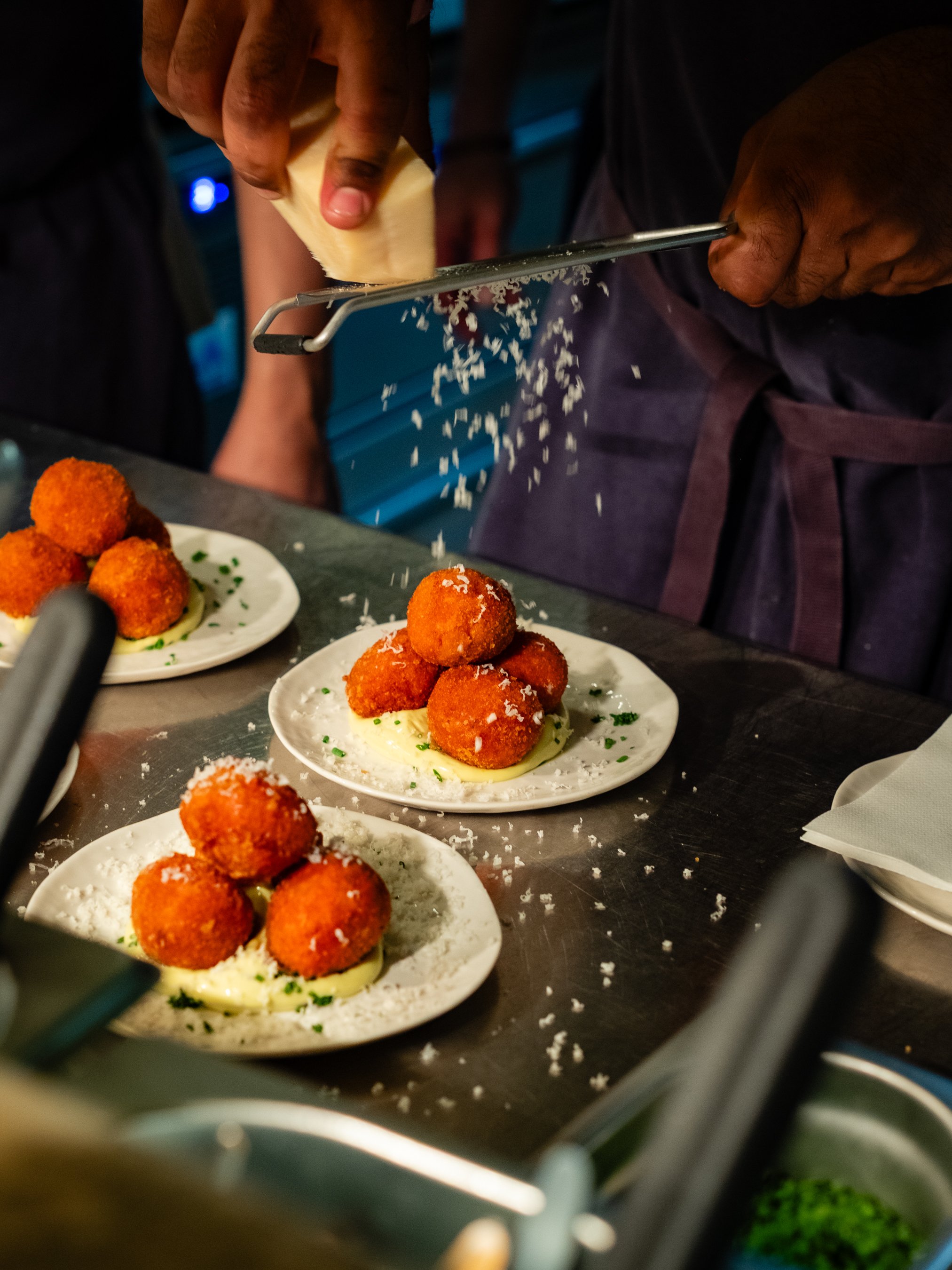 A person grating cheese onto a plate of fried croquettes served on a bed of yellow sauce, with another plate of croquettes nearby on a busy restaurant table.
