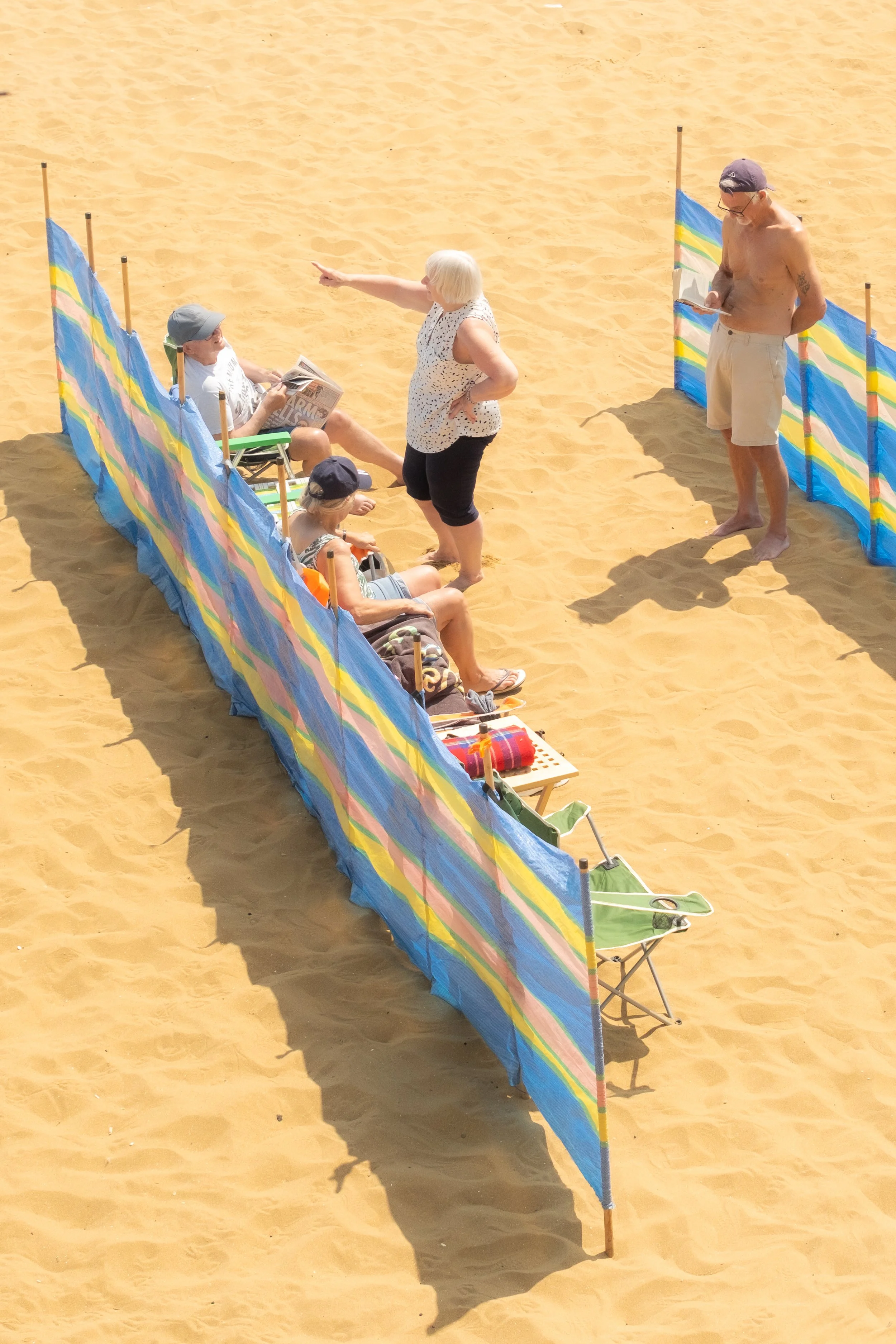 People on a sandy beach behind a colorful striped windbreak, some sitting and reading, one woman standing and pointing, one man standing shirtless, wearing sunglasses and a cap, looking at his phone.