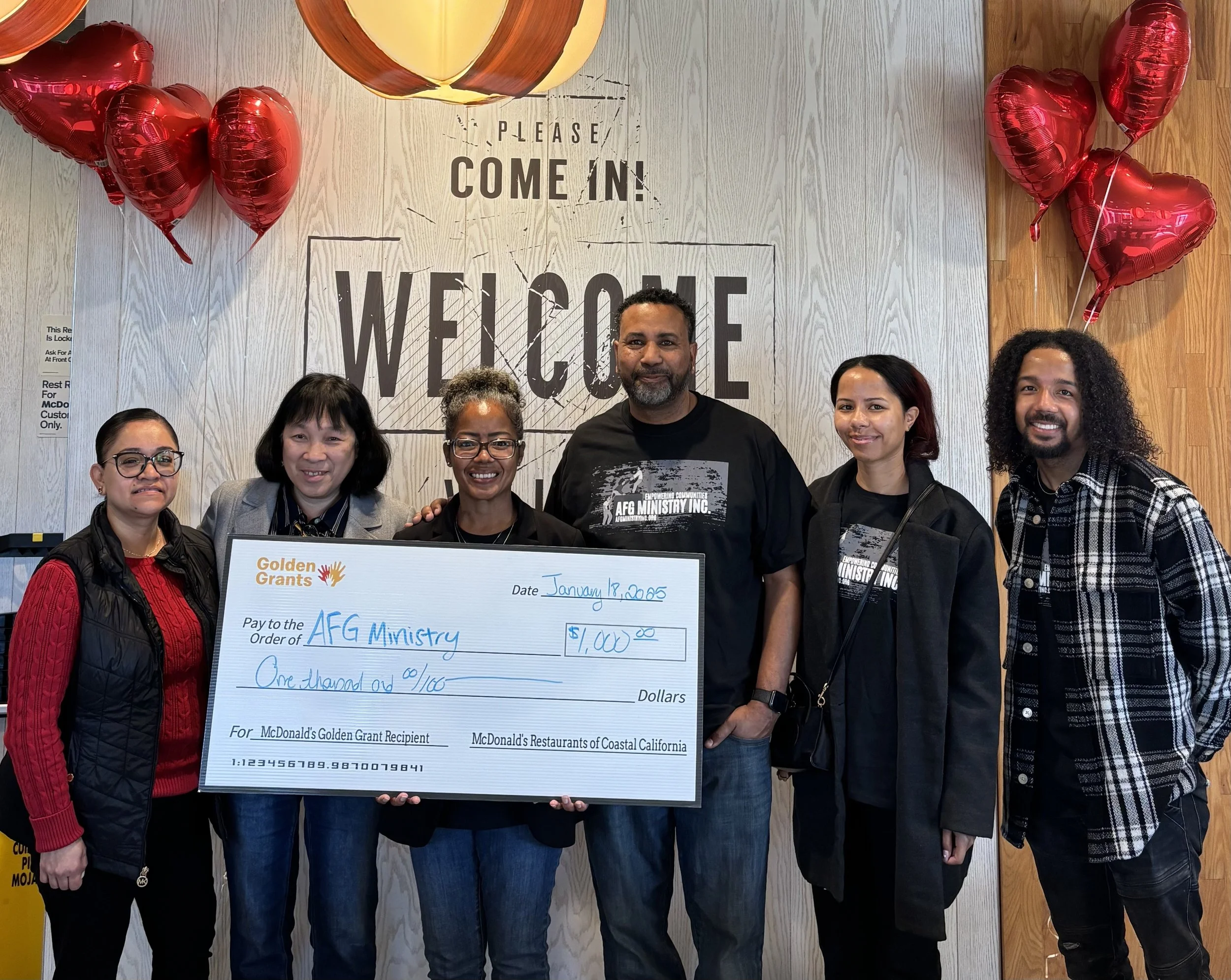 A group of six people standing in front of a wall with a sign that says 'Please Come In! Welcome'. They are holding a large check for $1,000 made out to AFG Ministry, dated January 18, 2023, from McDonald's Golden Grant. The wall has red heart-shaped balloons on either side and a wooden panel. The group is smiling and dressed casually, with some wearing black t-shirts with white text.