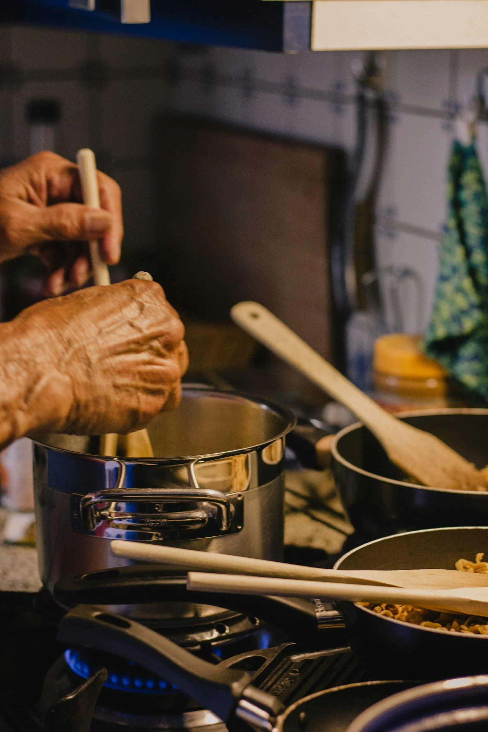 Close-up of elderly person's hands preparing food on a stovetop with pots and pans, a wooden spatula, and kitchen utensils.