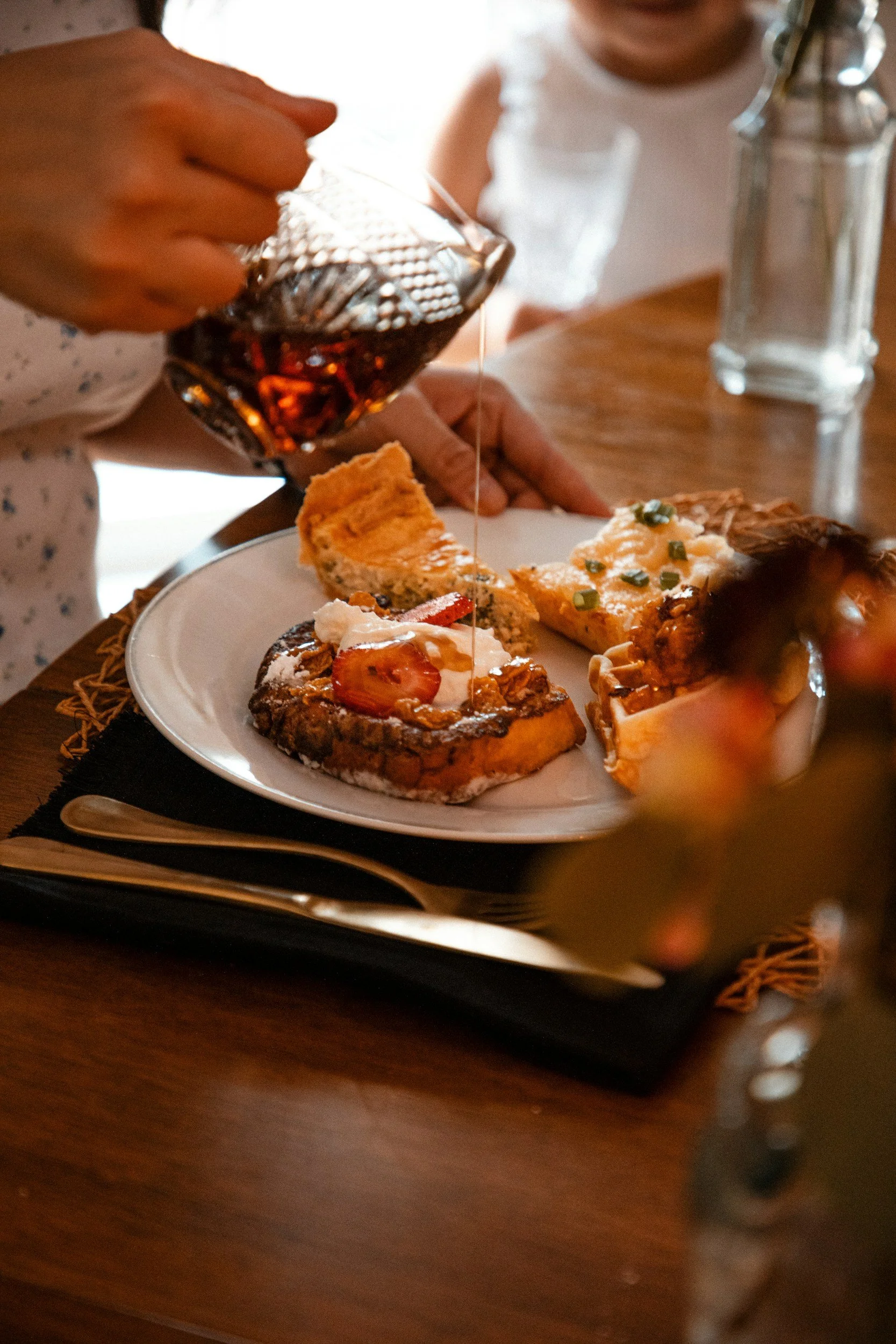 A plate of assorted desserts including cake slices topped with whipped cream and strawberries, and other pastry treats. A person is pouring syrup from a glass pitcher onto the desserts. The setting appears to be a dining table.