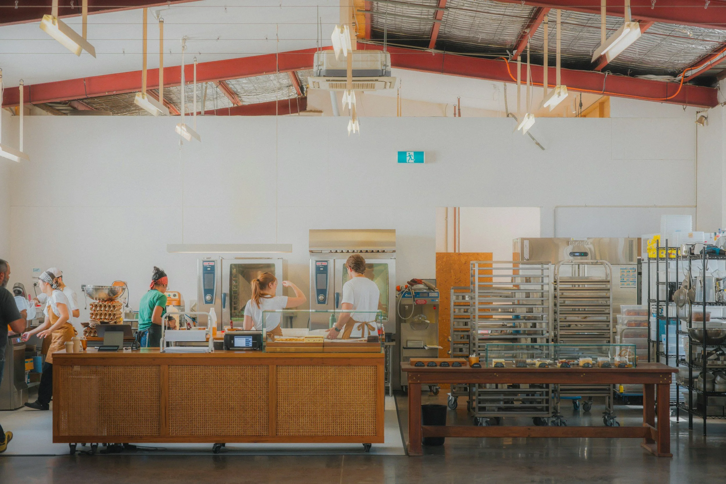 Employees working behind a counter in a commercial kitchen with industrial equipment and high ceiling.