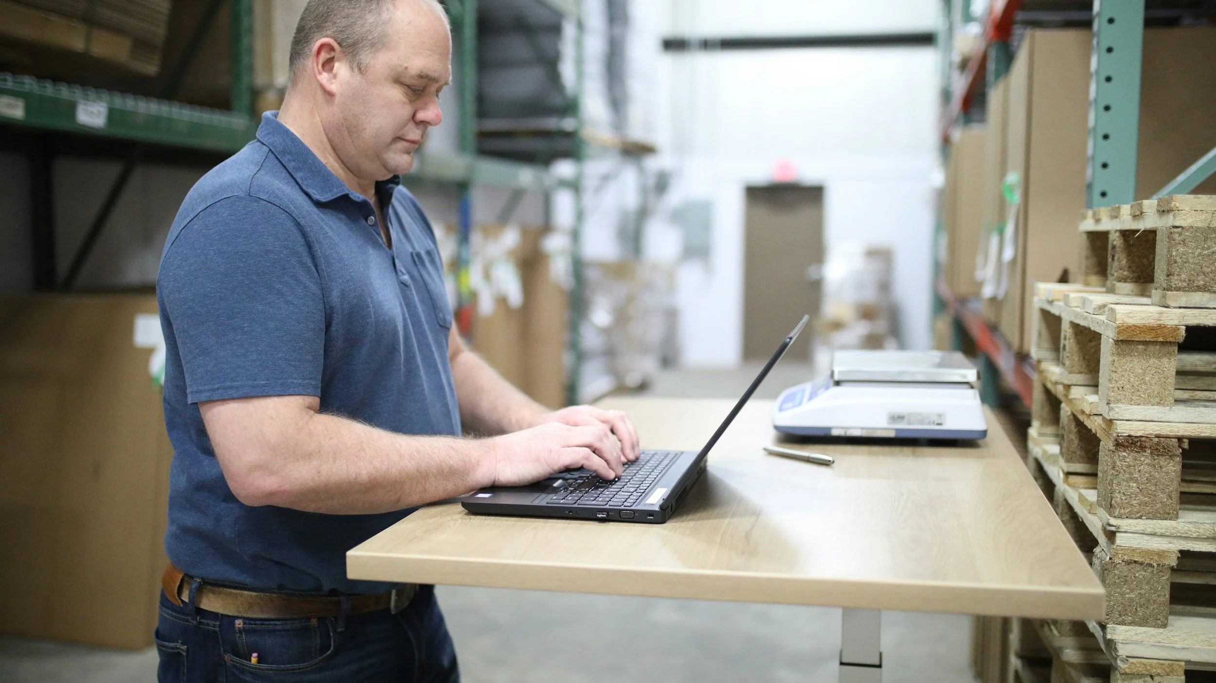 A man wearing a blue polo shirt working on a laptop at a warehouse or storage facility with shelves of boxes and wooden pallets.