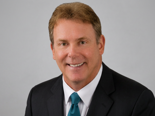 Professional headshot of a smiling Walt Carey owner of County Property Advisors in a black suit and teal tie against a gray background. Serving Eastern North Carolina for their property selling needs.