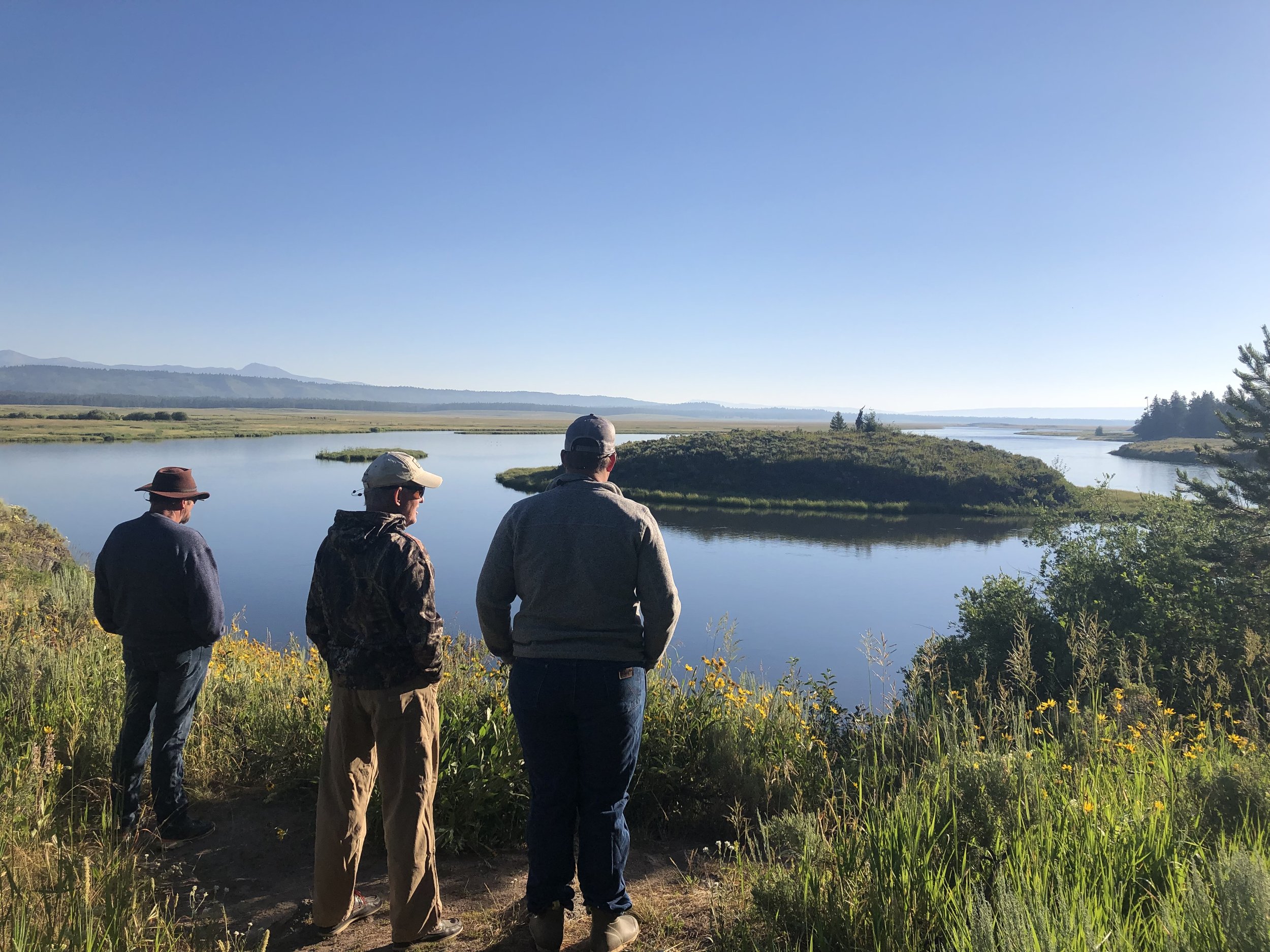 Three people stand by a river, looking at the scenic landscape with grassy banks, water, trees, and distant hills under a clear blue sky.