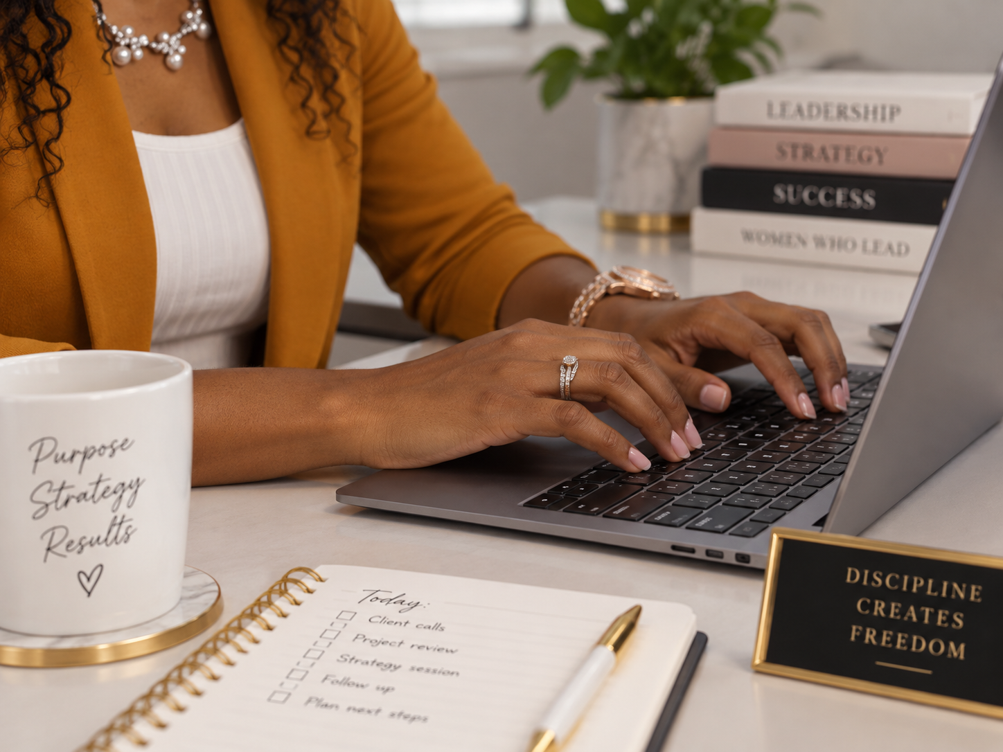 A woman working on a laptop at a desk with motivational books, a coffee mug, a notebook, and a framed quote. She is wearing jewelry and a mustard-colored blazer.