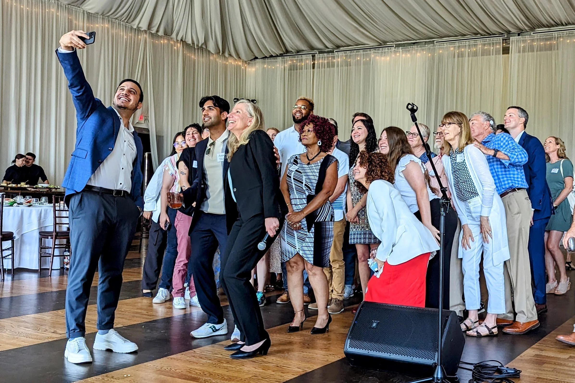 A group of people at an indoor celebration or event, with some taking selfies and smiling, and others chatting and enjoying the moment.