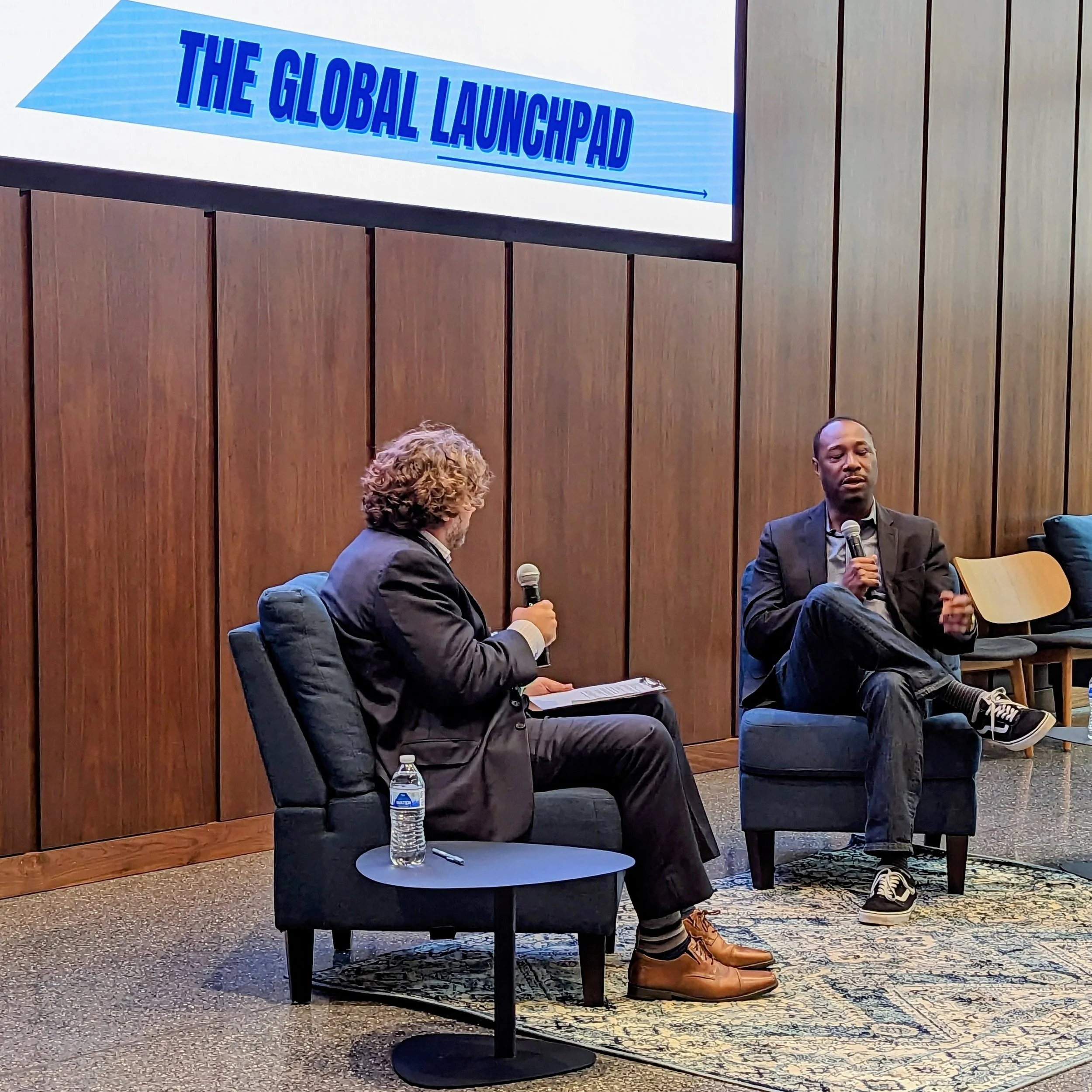 Panel with text 'The Global Launchpad' at an event, individuals sitting and speaking with microphones, water bottle on a small table, seen in a room with wooden wall panels.