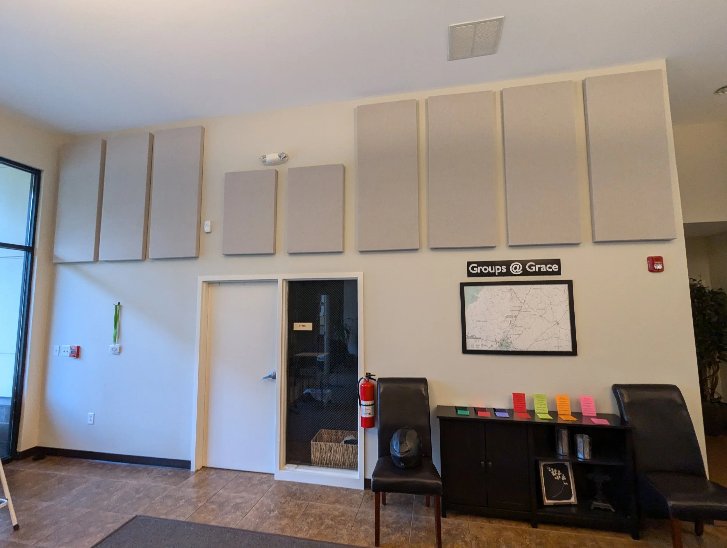A wall in a room with gray acoustic panels, a door with a window, a fire extinguisher, and a sign that says 'Groups @ Grace'. There are chairs and a table with colorful sticky notes on it.
