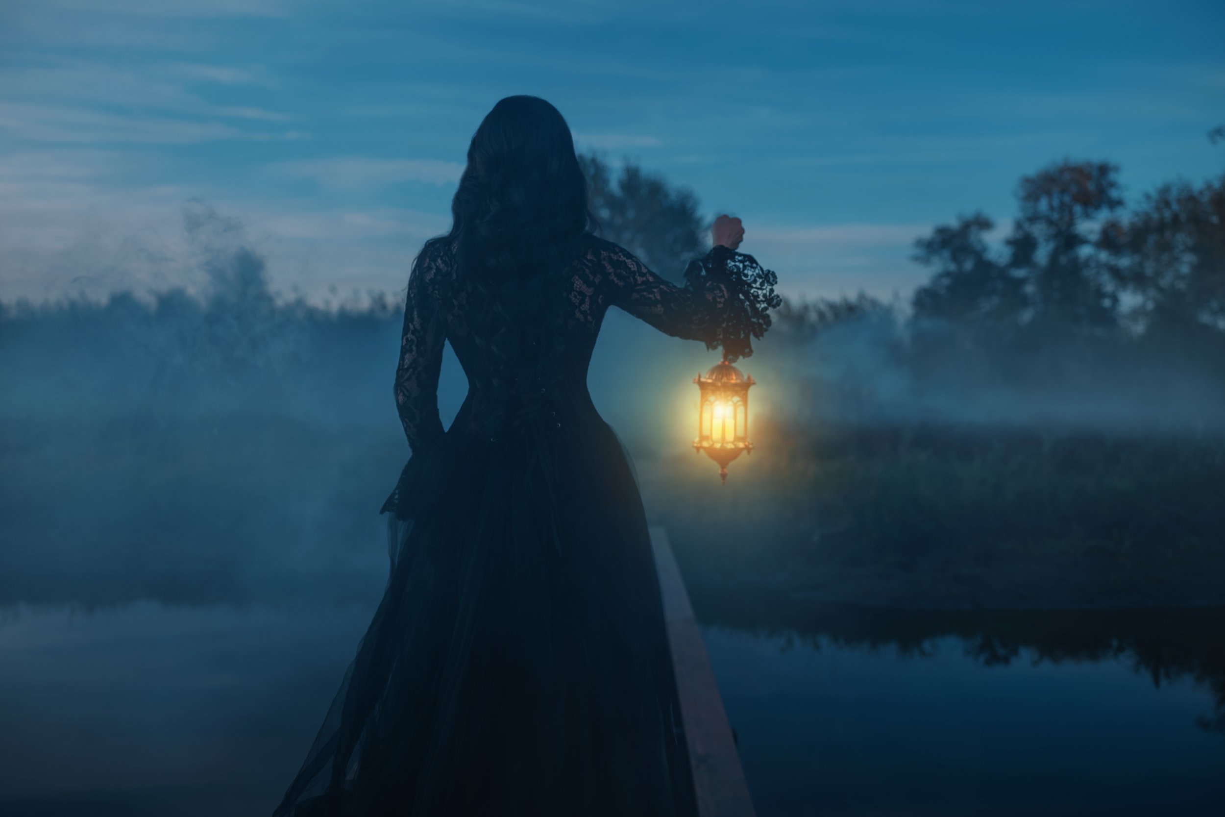 A woman in a black lace dress holding a glowing lantern outdoors during dusk or dawn, with trees in the background and fog near a body of water.