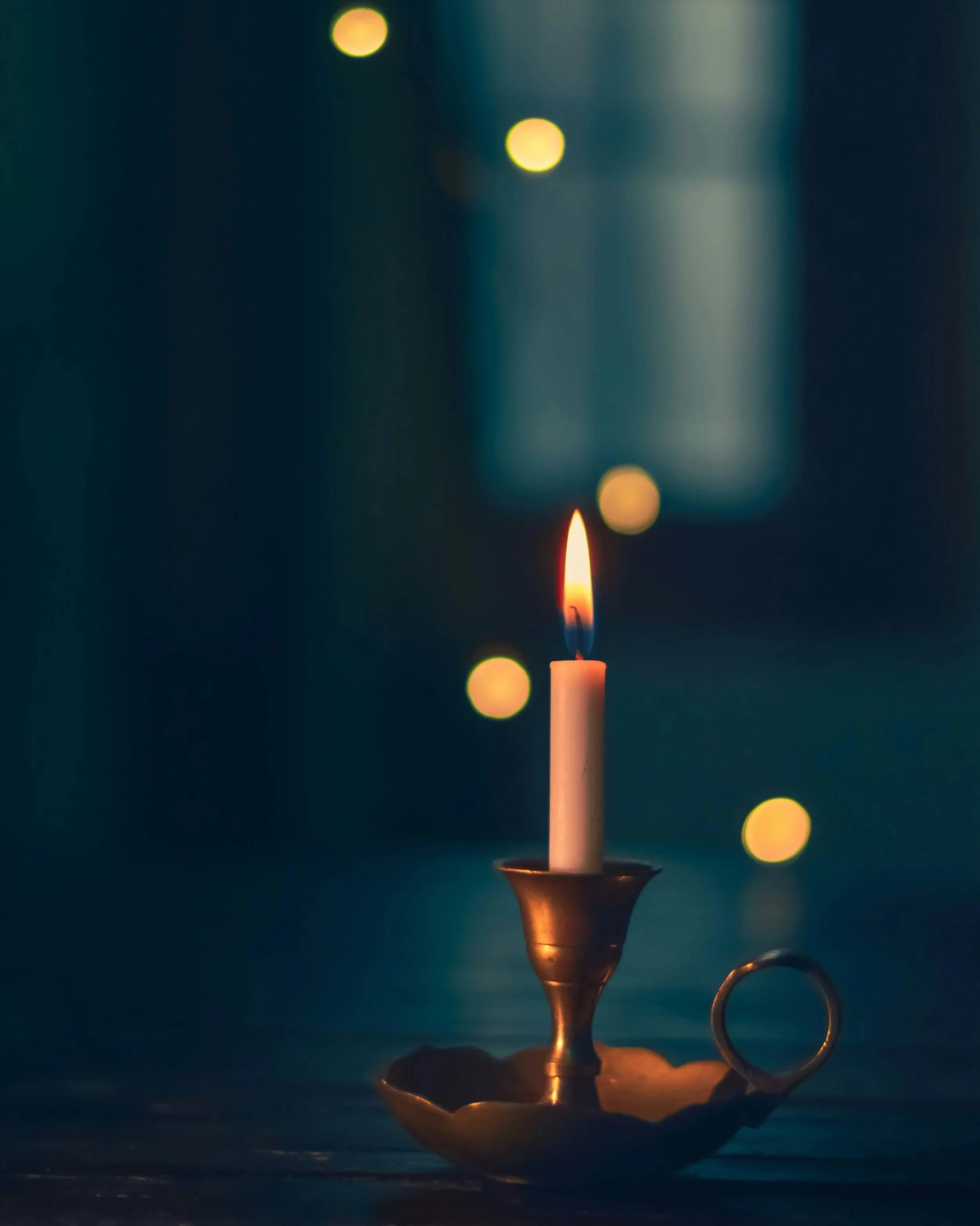 A lit candle in a candle holder with blurred circular yellow and green lights in the background.