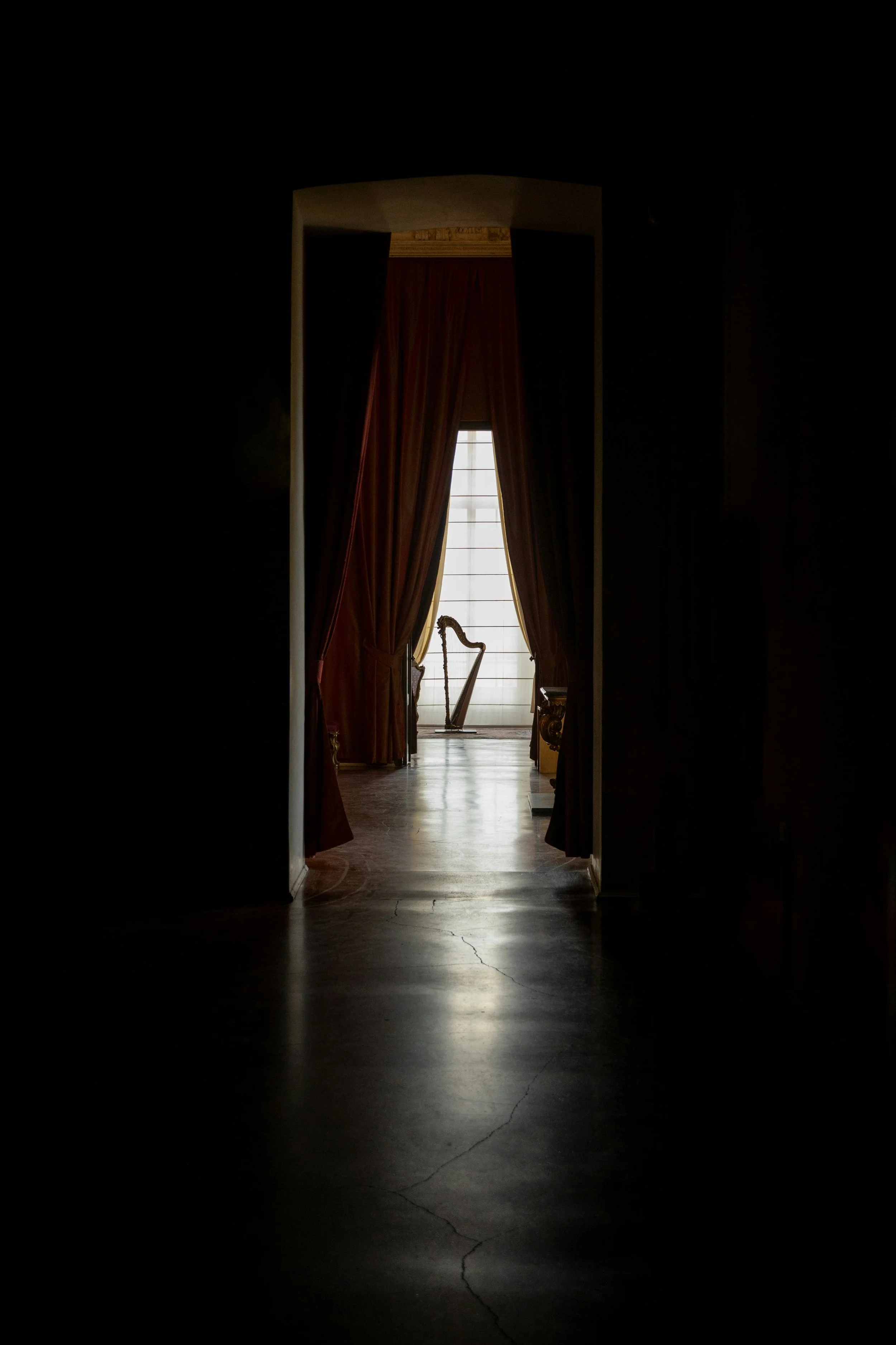 Dimly lit hallway leading to a room with large window and red curtains, with antique furniture and hardwood floors.