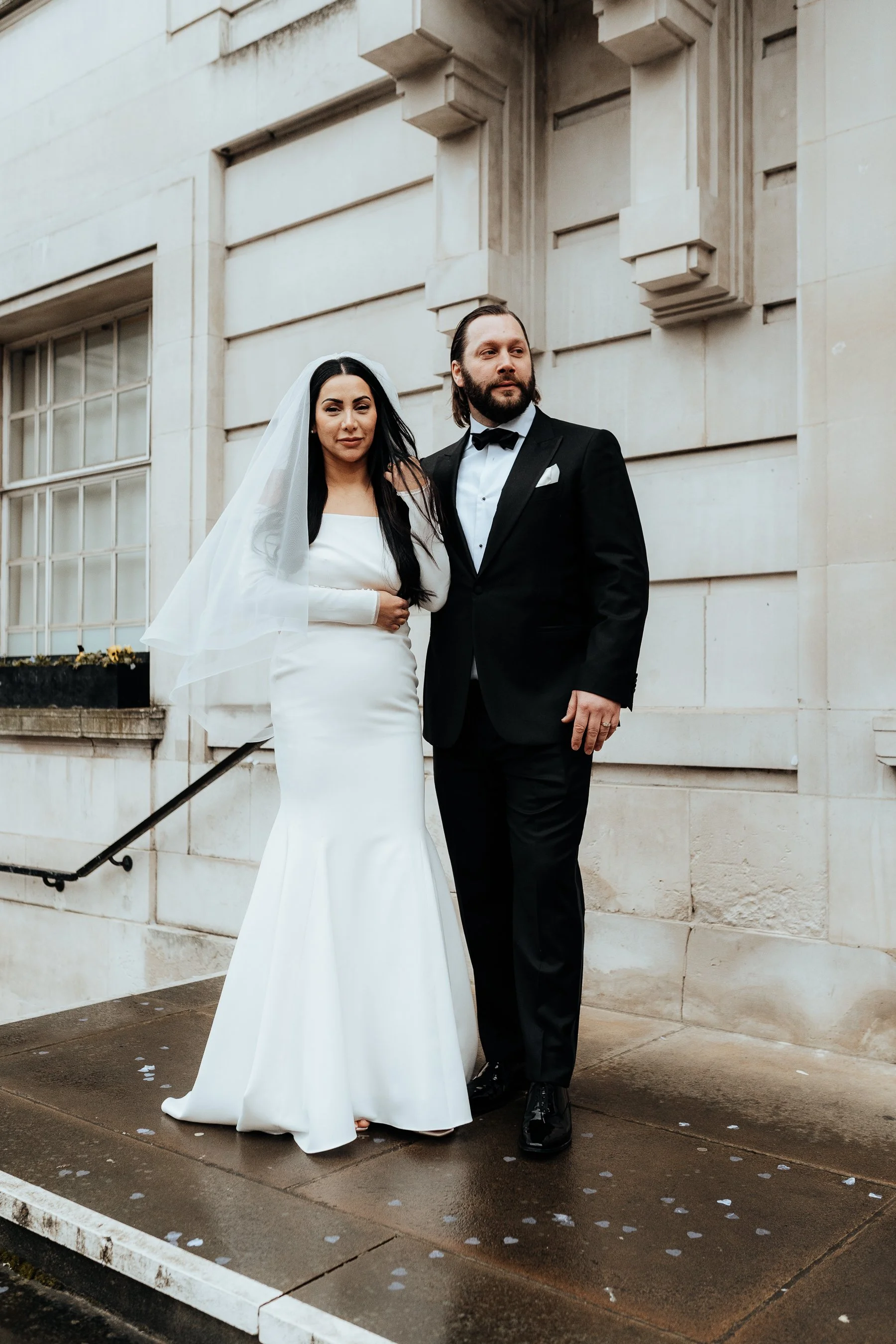 A bride and groom standing together on a wet sidewalk outside a stone building. The bride is wearing a white wedding dress with long sleeves and a veil, with long dark hair, and the groom is in a black tuxedo with a bow tie. They are looking in oppos