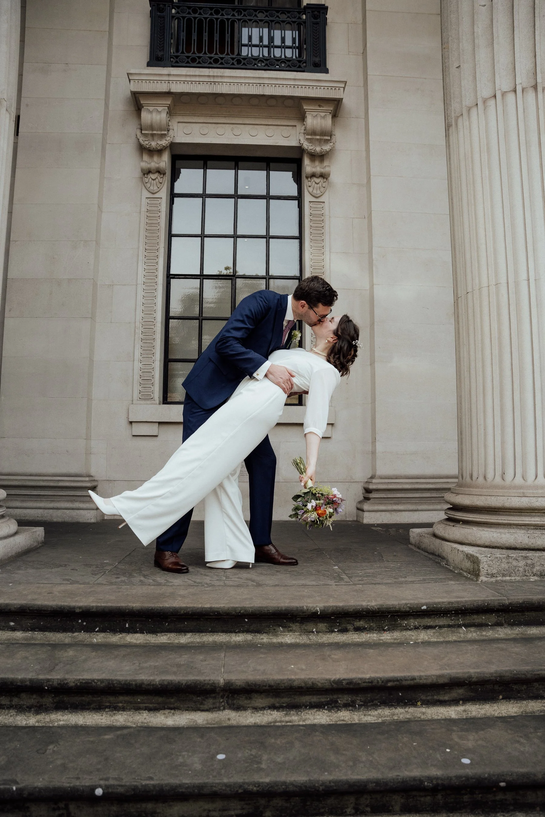 A newlywed couple stands on stone steps outside a large building with classical columns and a decorative window. The man, in a navy suit and glasses, dips the woman, dressed in a white wedding gown, for a kiss. She holds a bouquet of flowers.