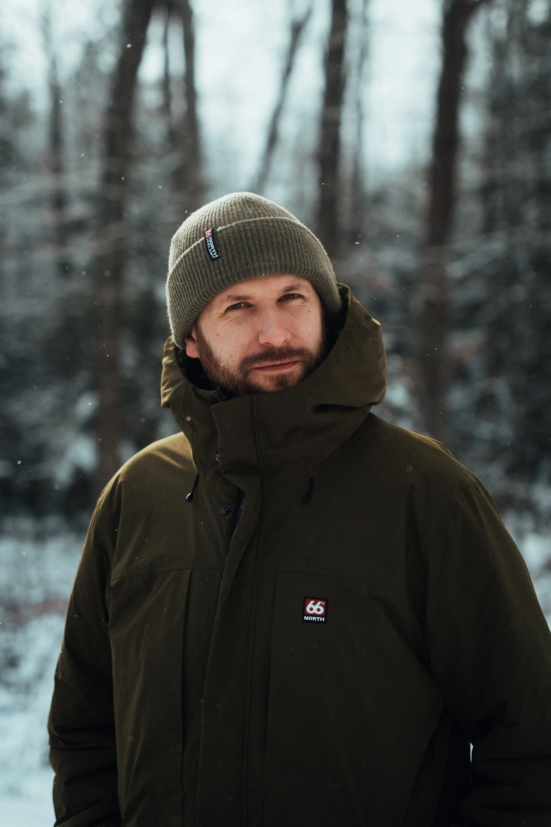 A man wearing a brown beanie and a dark green winter jacket with a '66 North' logo on it, standing in a snowy forest with tall, leafless trees in the background.