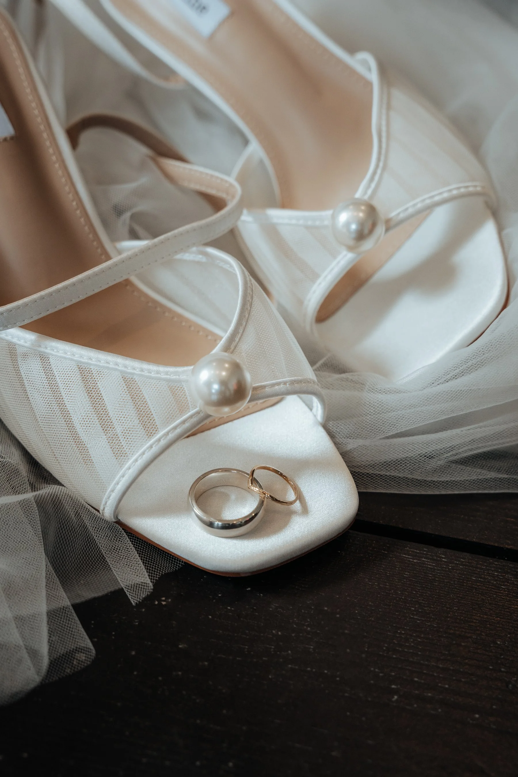 Close-up of wedding accessories including a pair of white bridal shoes with pearl embellishments, wedding rings, and soft tulle fabric on a dark wooden surface.