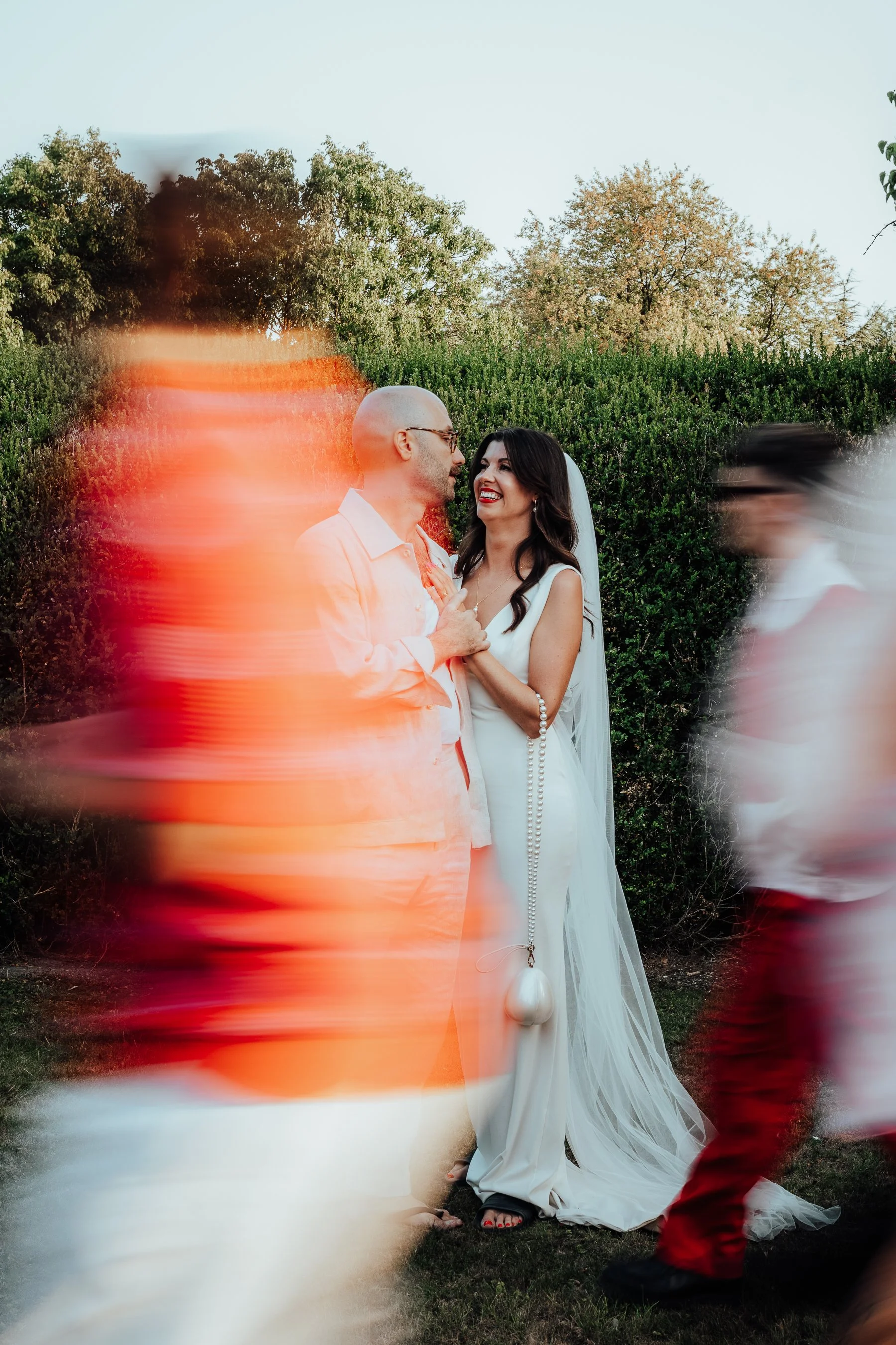 A bride and groom smiling at each other during their outdoor wedding ceremony, with blurred guests walking by in the foreground.