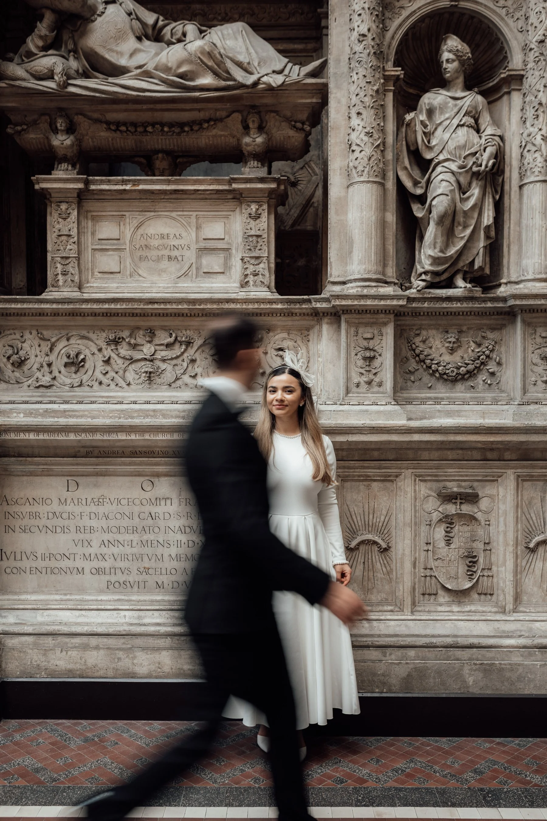 A bride in a white wedding dress and a large decorative headpiece stands in front of an ornate marble monument with sculptures and inscriptions. A groom in a dark suit is walking past in front of her, blurred due to motion.