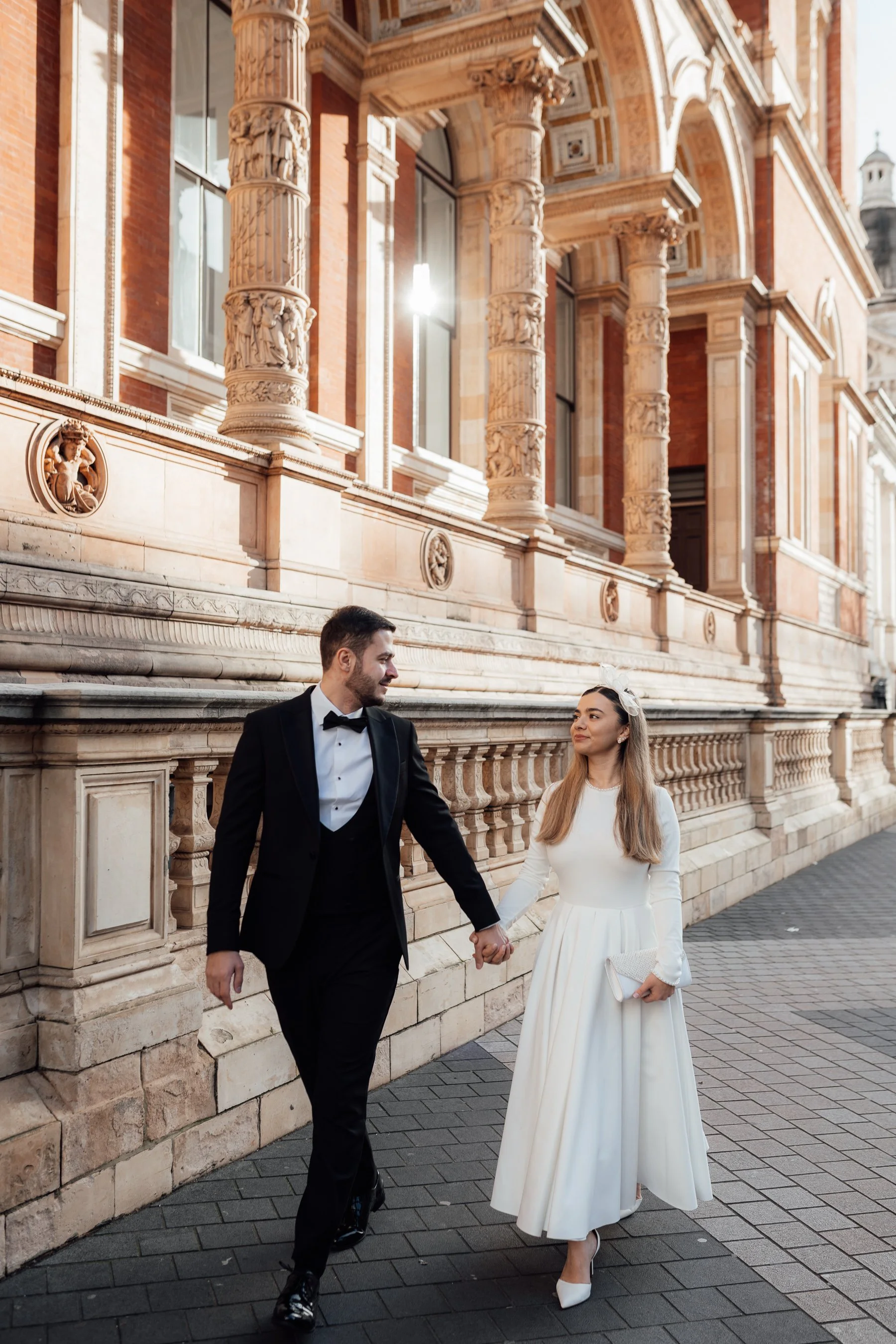 A bride and groom dressed in wedding attire holding hands, walking along a historic building with ornate columns and carvings, sunlight shining through a window.