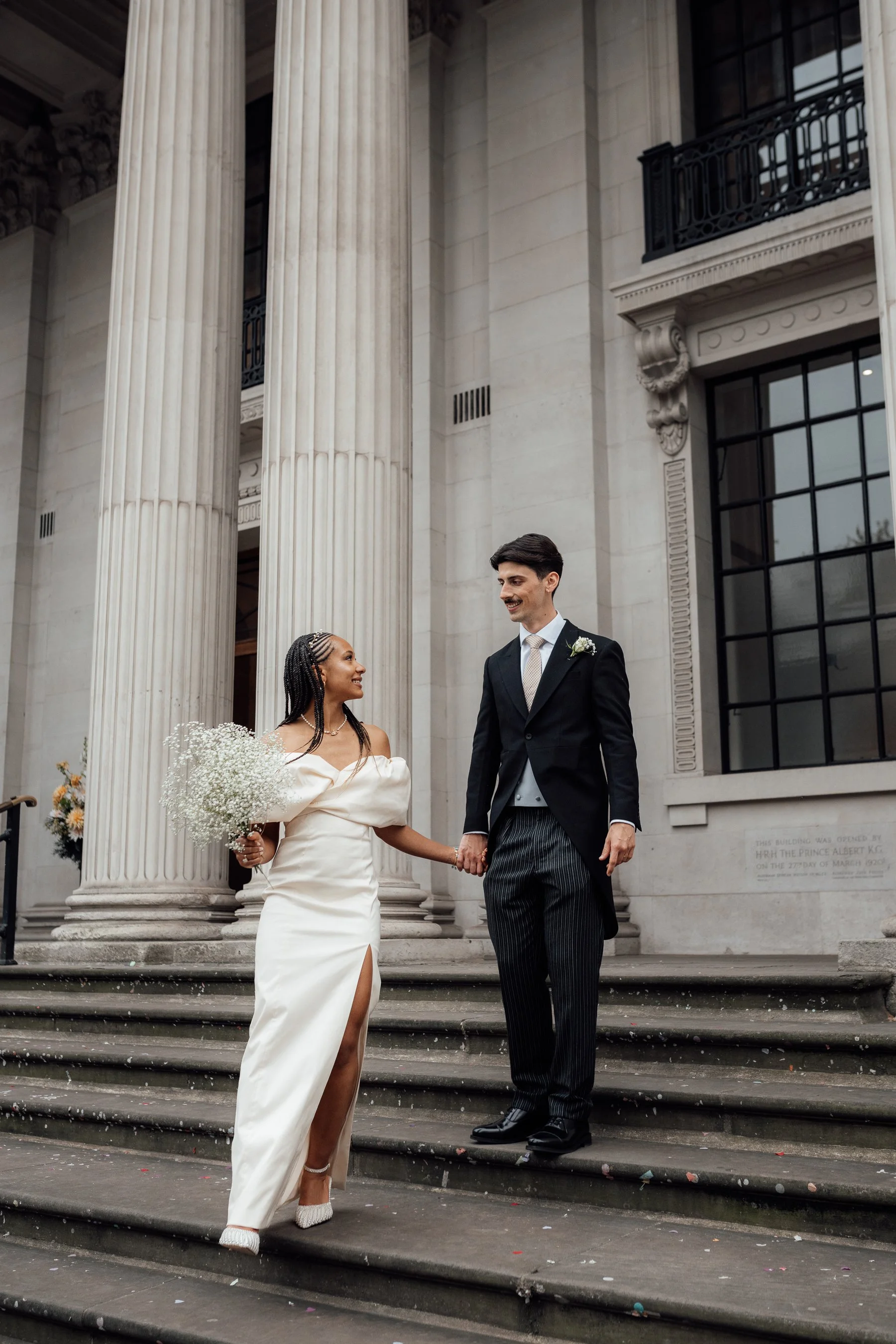 A bride in a white wedding dress holding a bouquet, and a groom in a black suit with a boutonniere, stand hand in hand on the steps of a historic building with large columns and decorative architectural details.