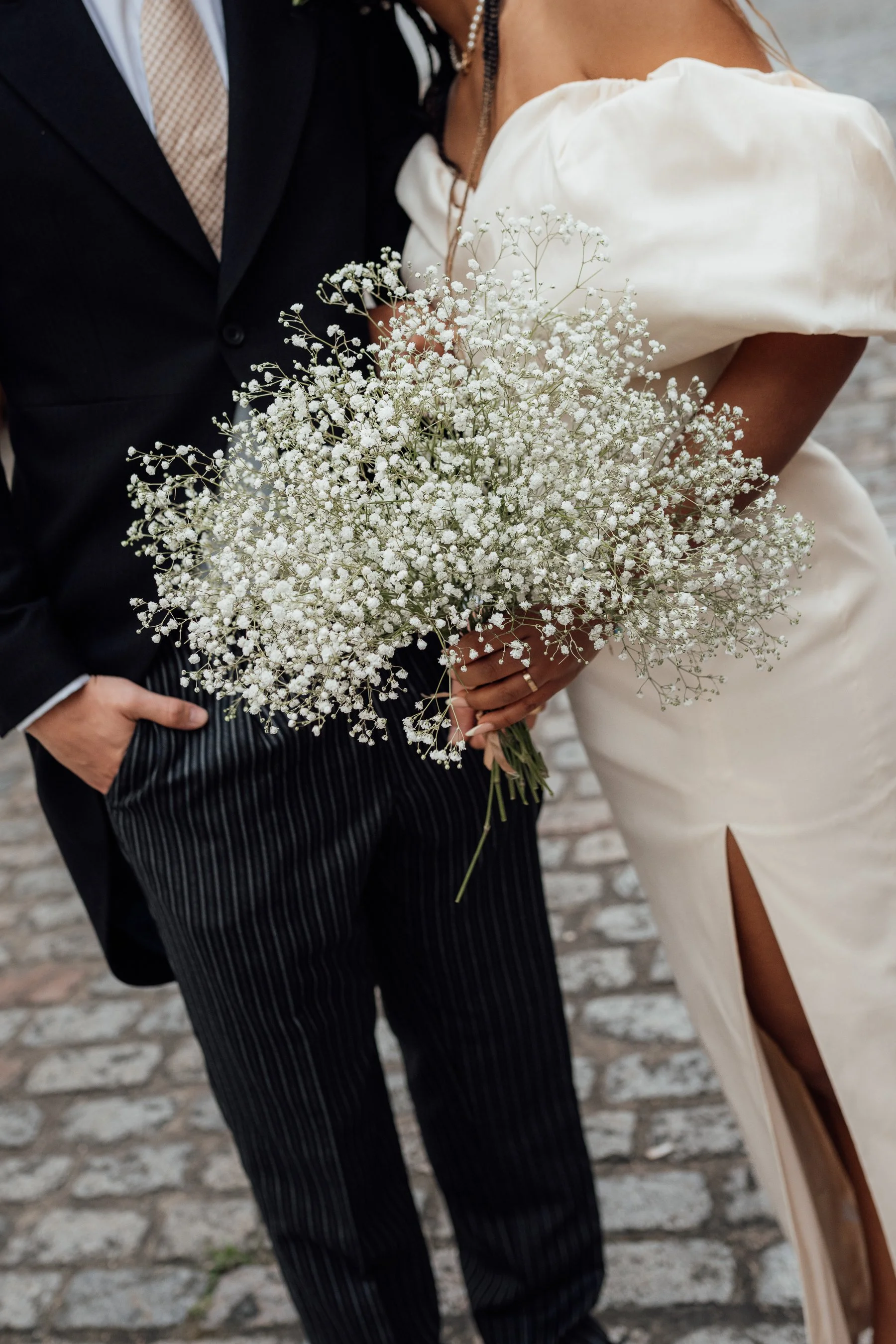 Close-up of a bride and groom standing together, with the bride holding a bouquet of white baby's breath flowers. The groom is wearing a dark suit, and the bride is wearing a white dress with puffed sleeves.