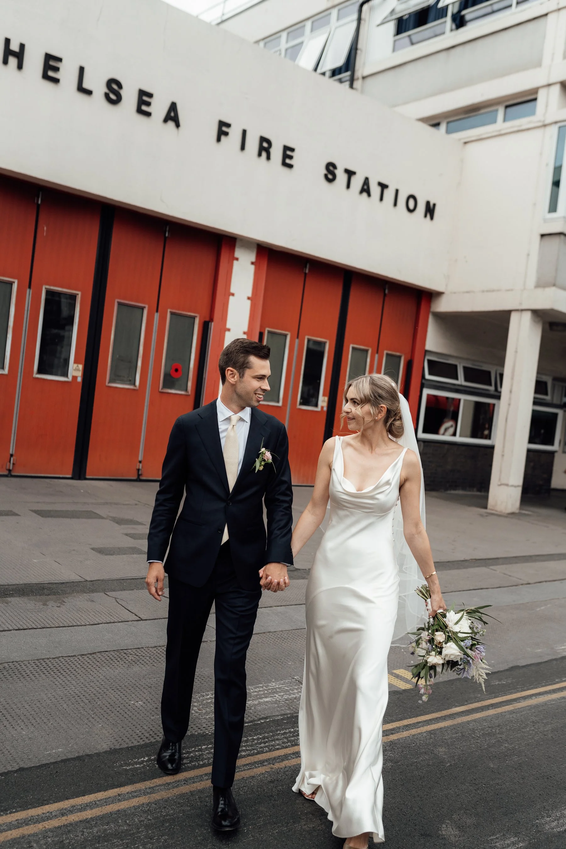 A newlywed couple walking hand-in-hand outside a fire station, with the groom in a dark suit and the bride in a white wedding dress, holding a bouquet of flowers.