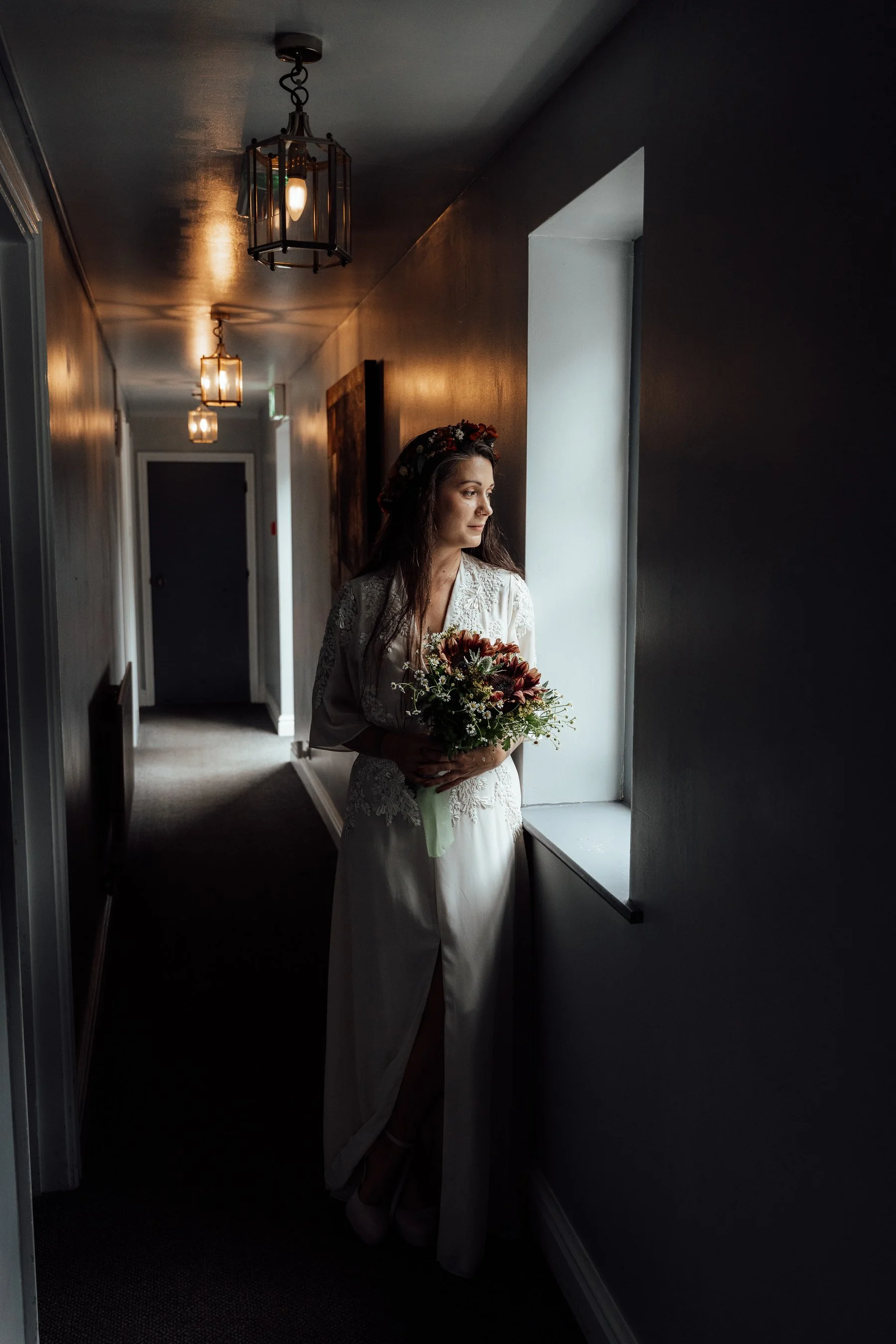 A bride in a wedding dress holding a bouquet of flowers, standing by a window in a dimly lit hallway with modern lighting fixtures.