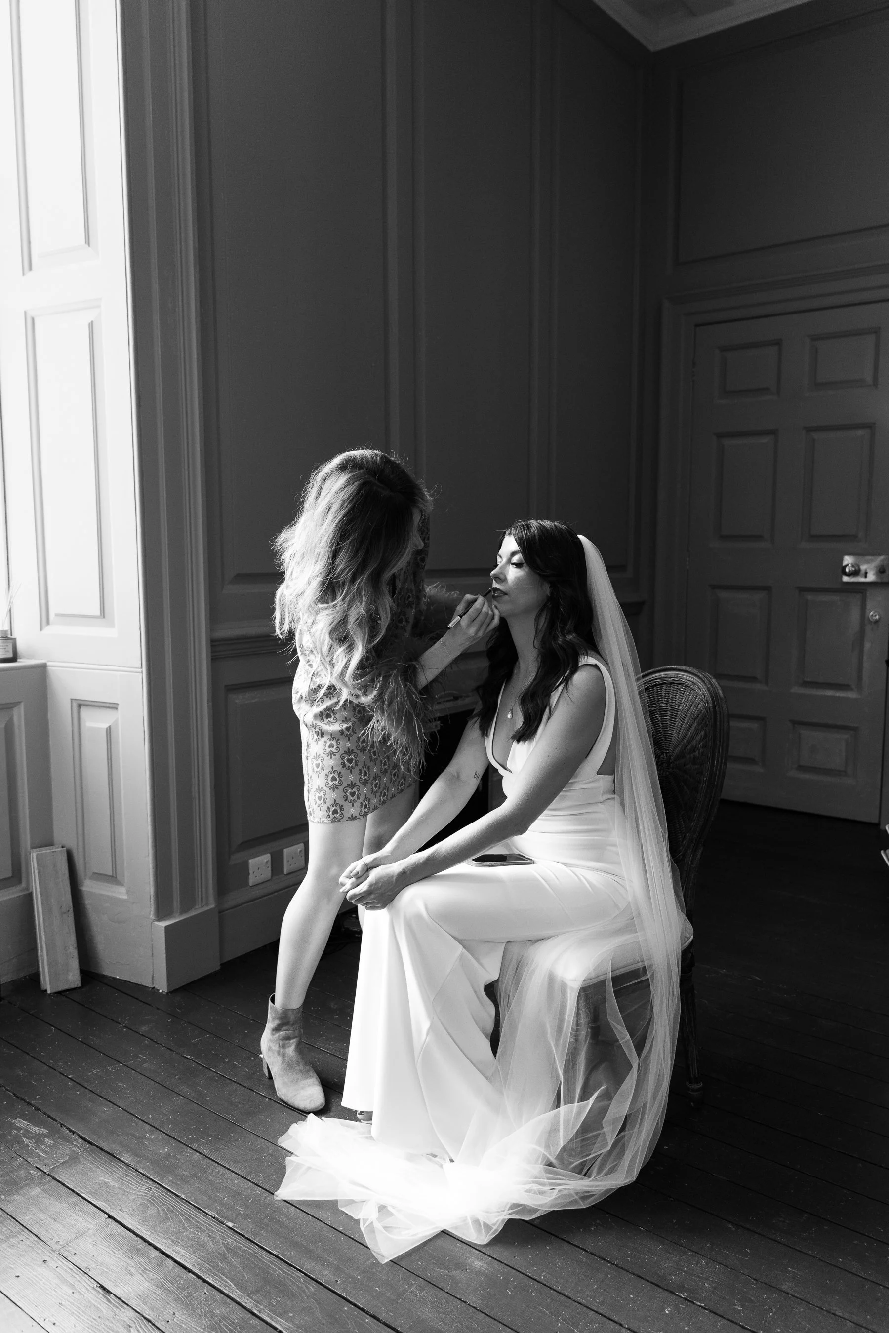 A bride with a veil sitting on a chair, having lipstick touched up by a woman standing in front of her. The bride wears a wedding dress with tulle on the skirt, and the woman touching up her lipstick has long, wavy hair and is wearing a lace dress. T