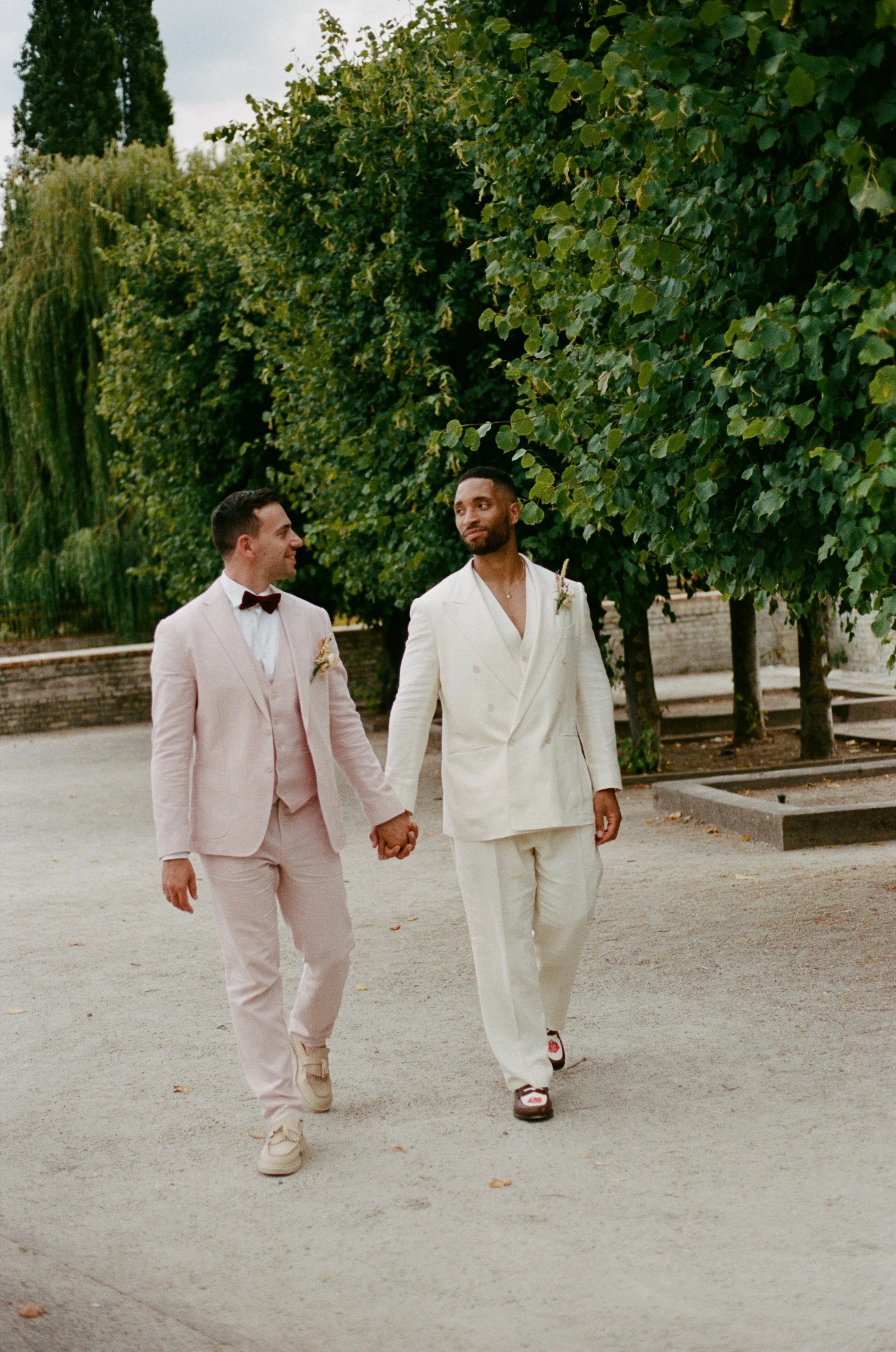 Two grooms in white suits are holding hands and walking outside in a park, surrounded by green trees.