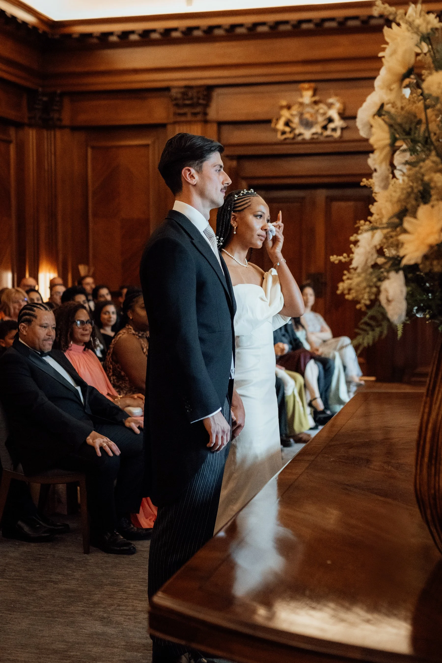 A couple, standing side by side, in formal attire during a wedding ceremony in a wood-paneled room with a floral arrangement and an audience seated behind them.