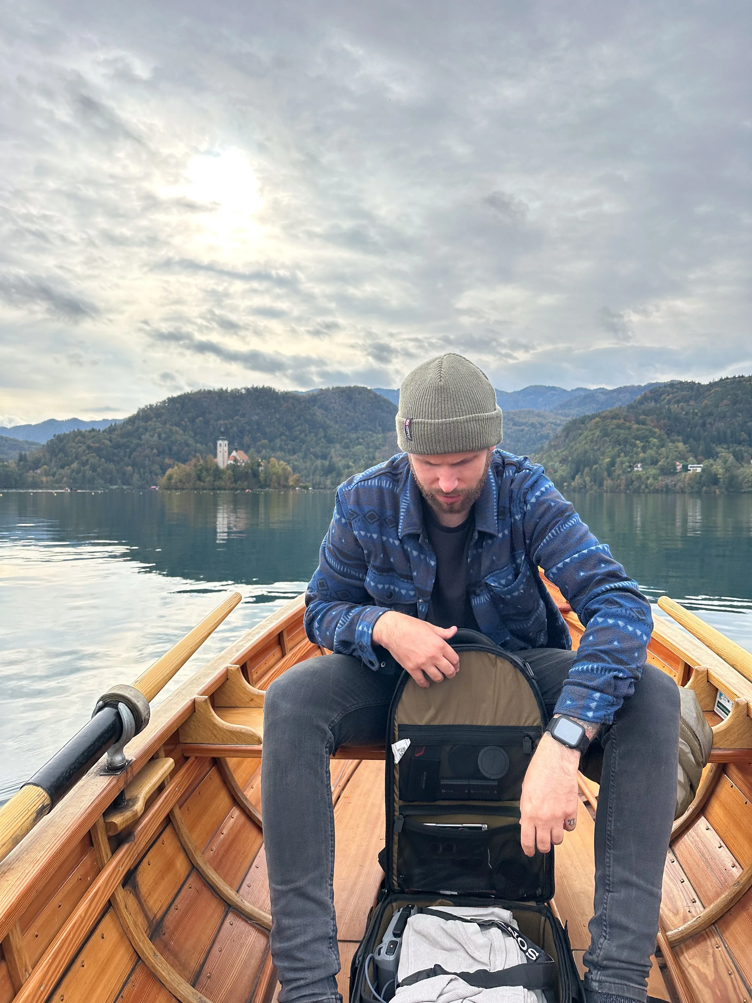 A man in a gray beanie and blue jacket sitting in a wooden rowboat on a lake with mountains and a cloudy sky in the background.