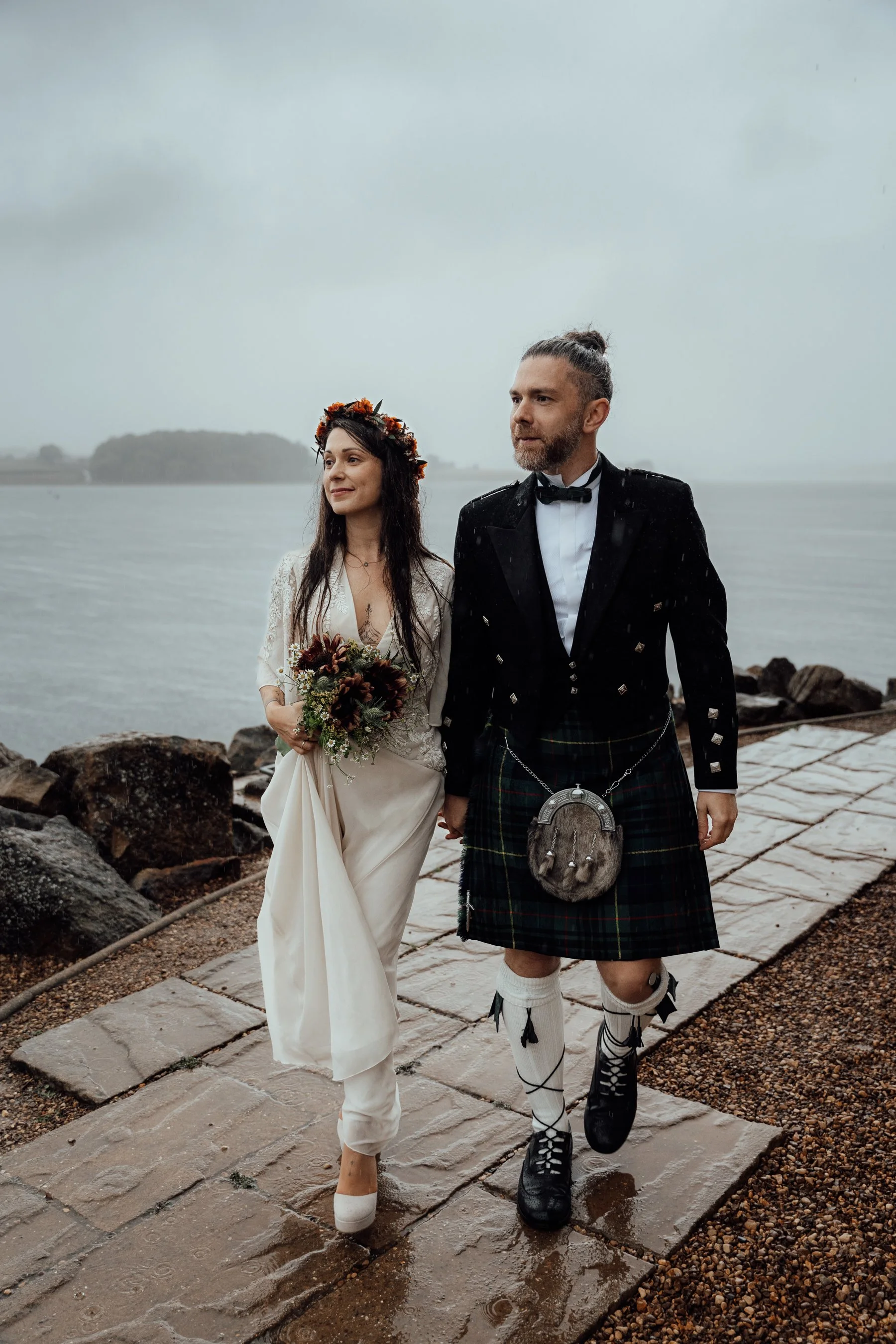 A couple dressed in wedding attire walking by the water on a rainy day, with the woman holding a bouquet and wearing a floral crown, and the man wearing a traditional Scottish kilt and sporran.