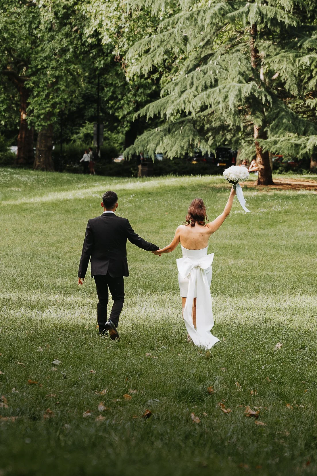 A newlywed couple walking hand in hand on a grassy lawn, with the bride holding a bouquet of white flowers raised in the air, surrounded by green trees in a park.