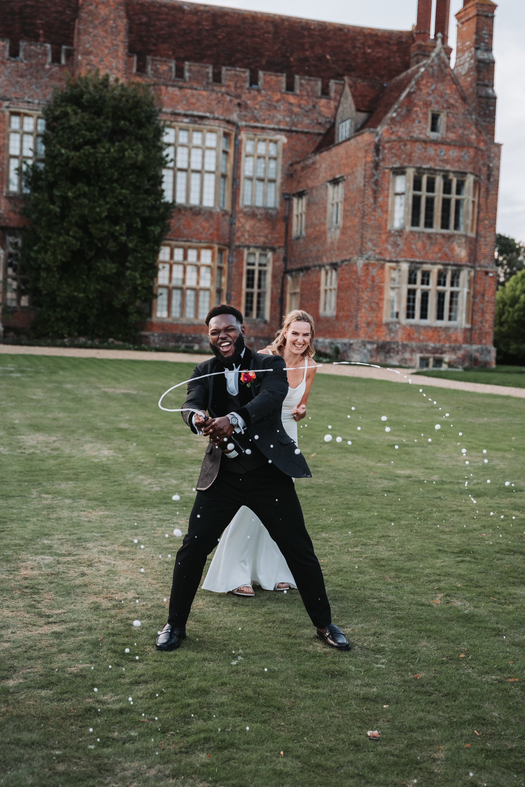 A newlywed couple celebrating outside on the lawn of a large brick castle-like building, with the groom firing a champagne bottle and the bride smiling behind him.