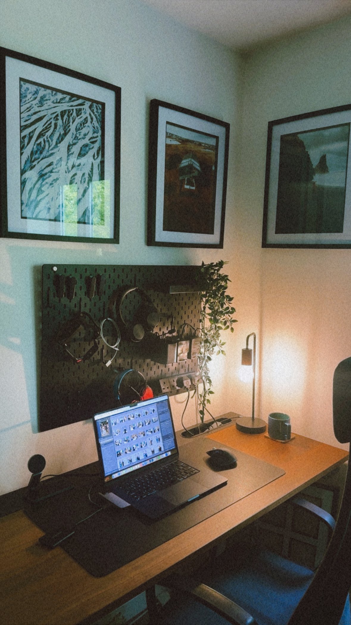 A home office workspace with a wooden desk, a laptop showing photo editing software, a mouse, headphones, and a small potted plant. There is a pegboard on the wall holding various items, a lamp with a warm glow, and wall art including three framed pictures of nature and landscapes.