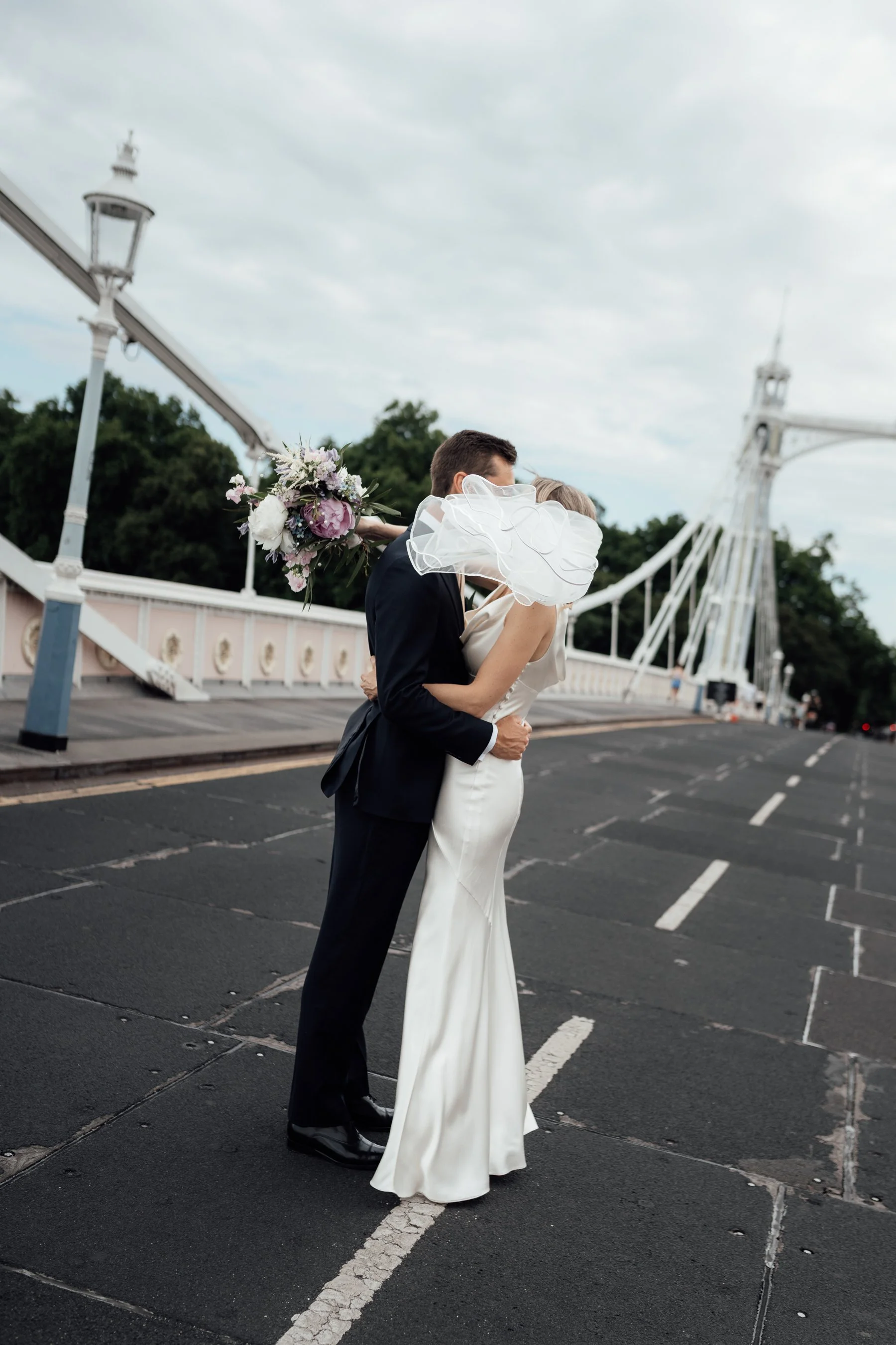 A bride and groom are embracing and kissing on a bridge with decorative railings, with the bride holding a bouquet of flowers, against a background of trees and an overcast sky.