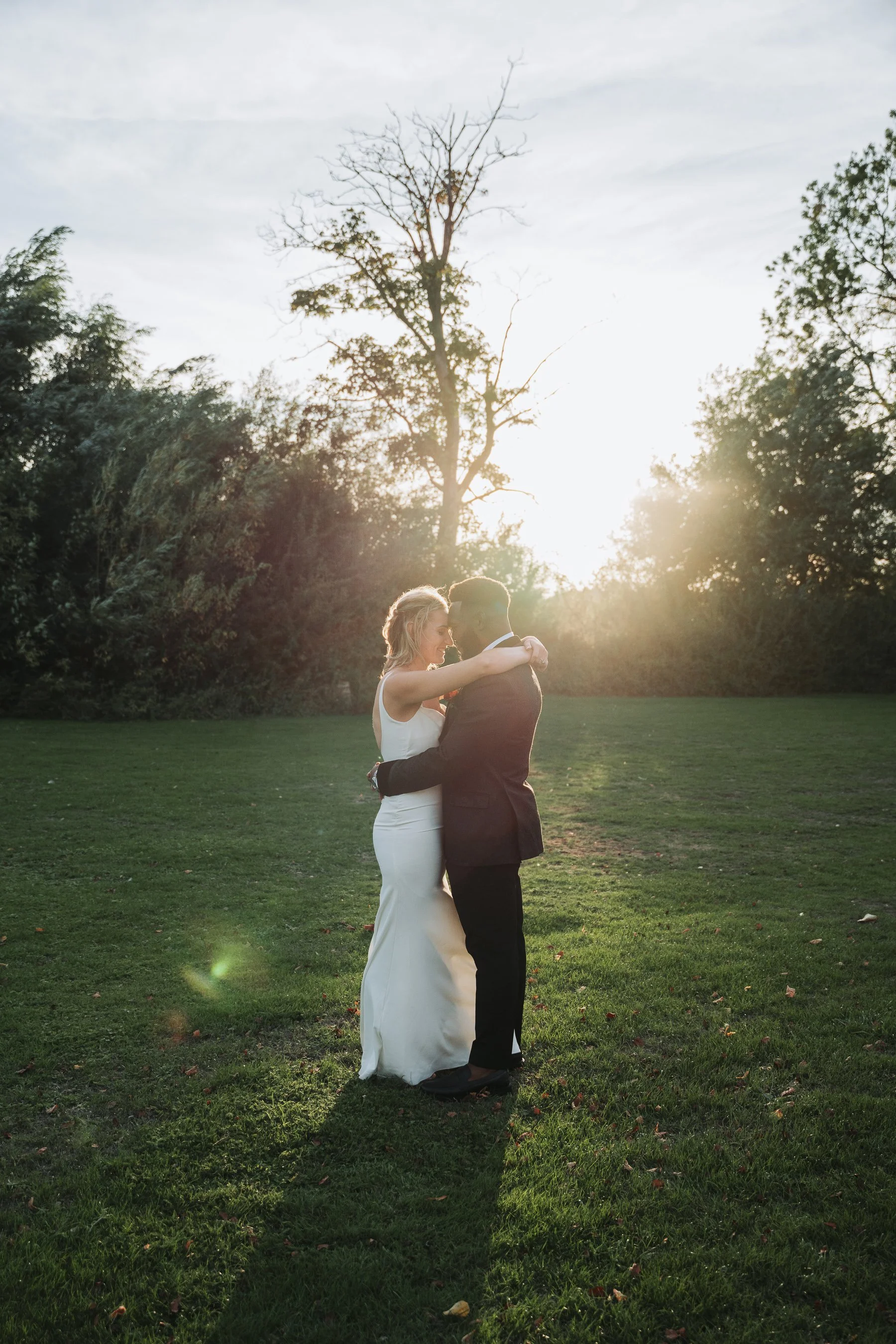A newlywed couple stands close together and shares an intimate moment outdoors during sunset, with trees in the background and a grassy field surrounding them.